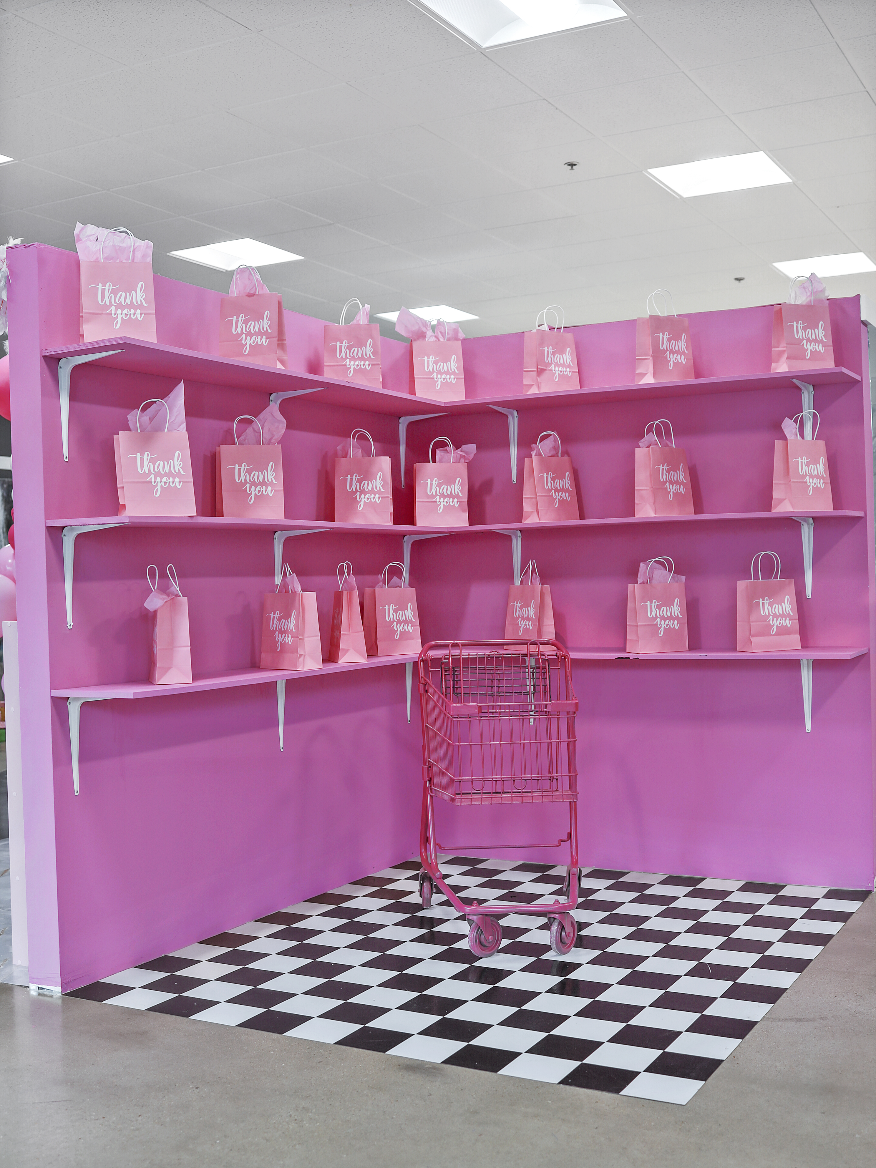 A vibrant pink shelving display filled with numerous matching pink bags labeled "thank you!" with a pink shopping cart on a black and white checkered floor in front.
