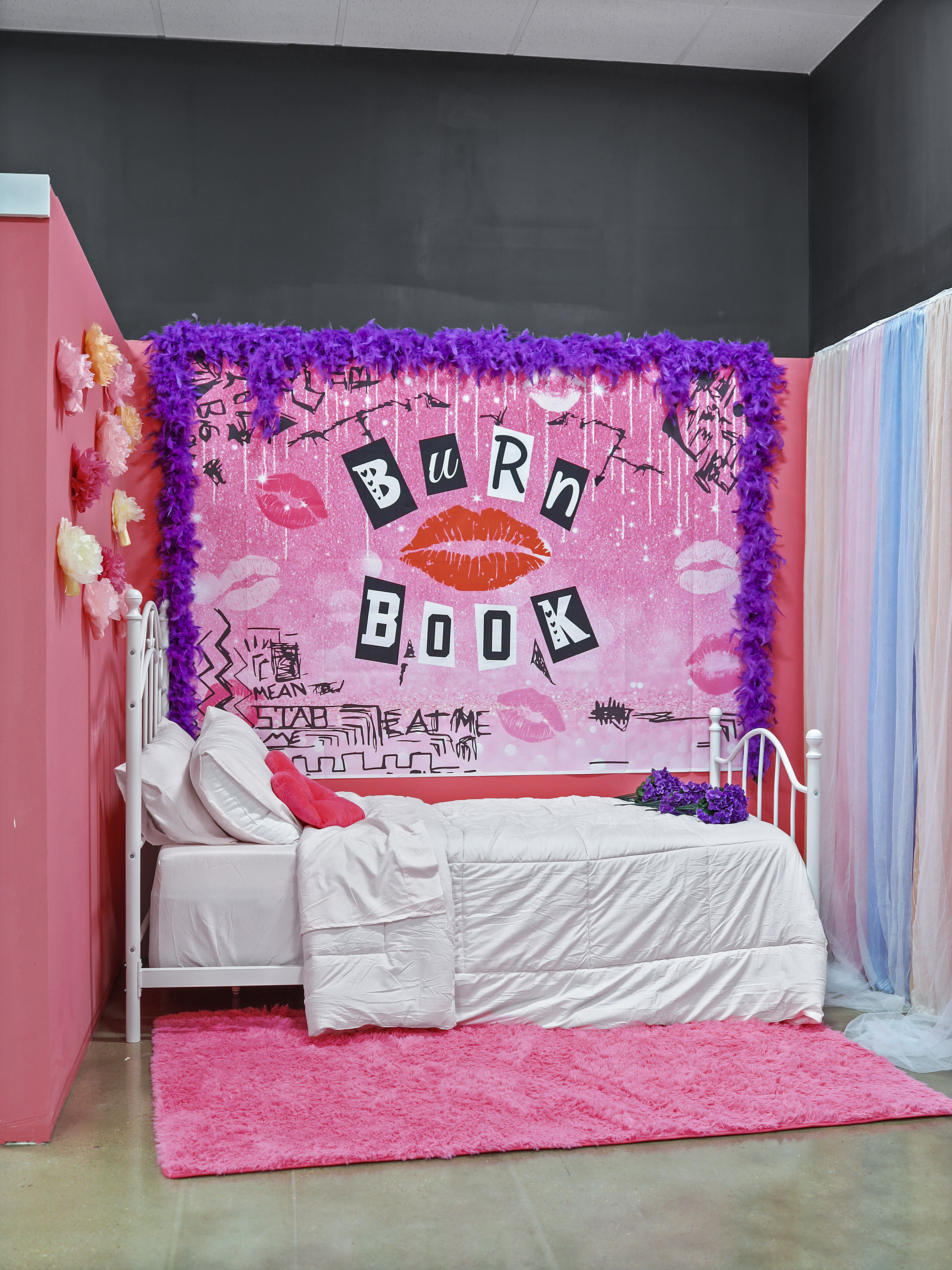 A vibrant bedroom setup with a white bed against a wall decorated with a pink "burn book" mural, surrounded by purple decorations and a pink rug.