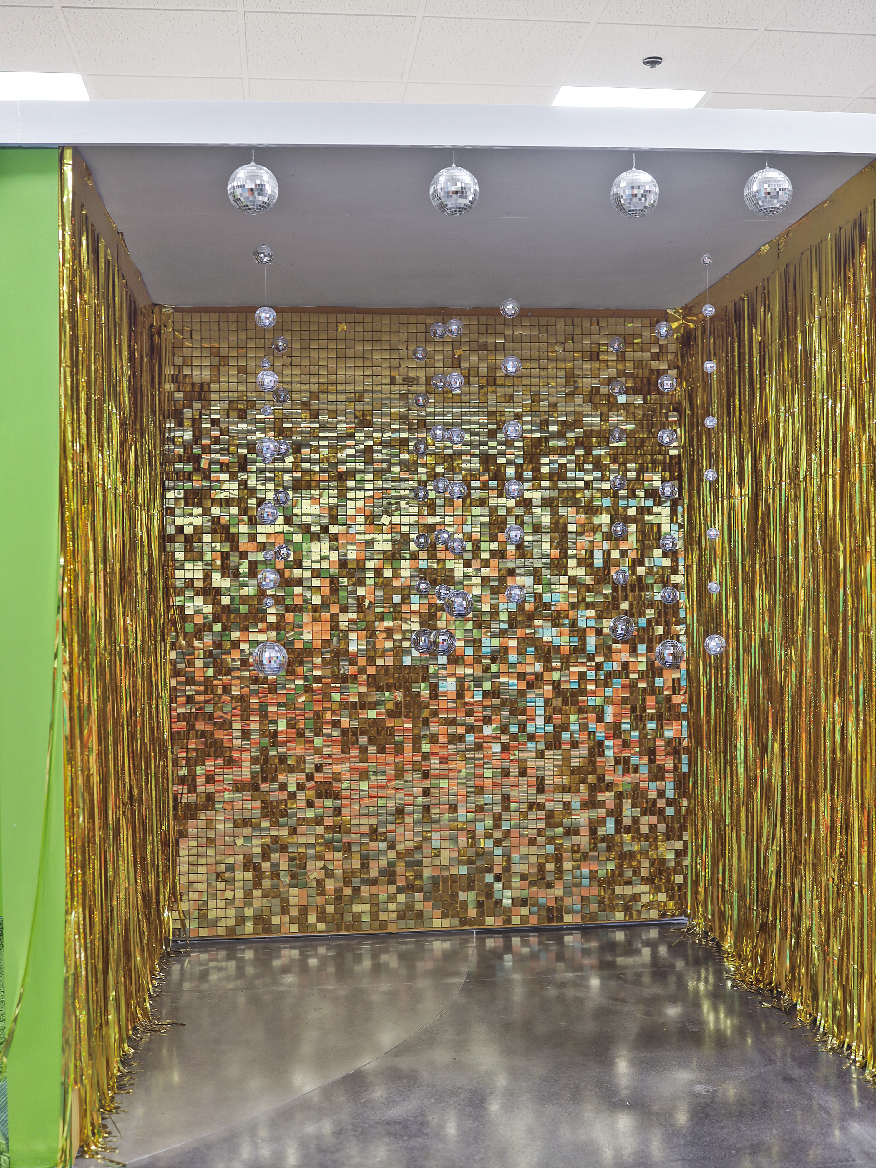 A vibrant art installation with a wall covered in a mosaic of shimmering gold squares and a curtain of gold streamers, flanked by disco balls hanging from the ceiling.