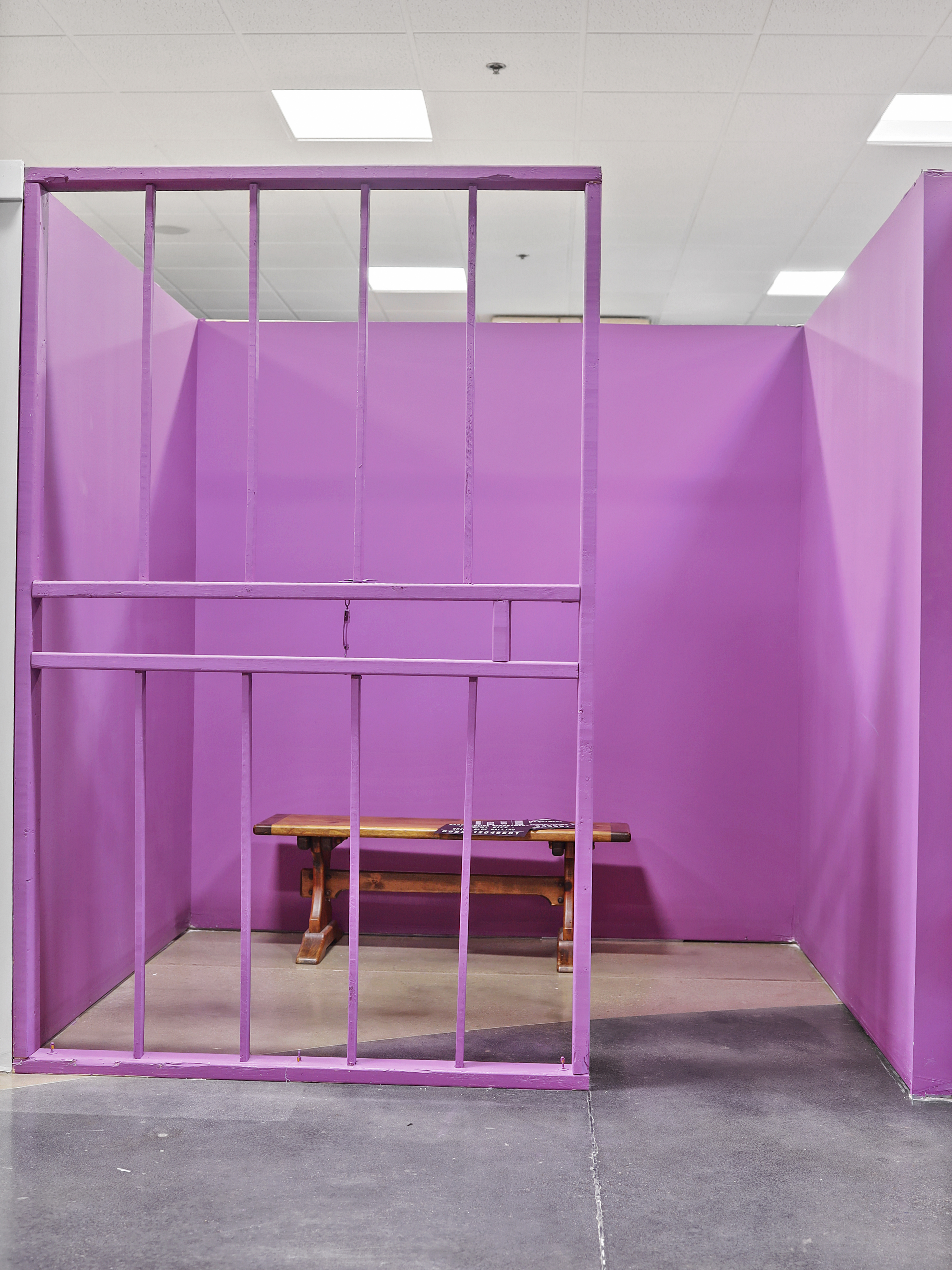 An indoor installation featuring a pink-painted wooden jail structure with bars at the front, containing a simple wooden bench against a pink background.
