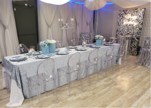 Elegant event table setup featuring a marble-patterned tablecloth with clear acrylic chairs. The table is set with silver chargers, white plates, and blue floral centerpieces. A shimmering backdrop adds
