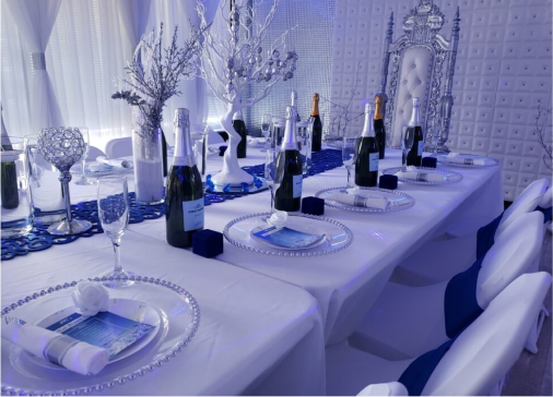 Elegantly set dining table on the homepage of a blue-themed event, featuring illuminated white trees, silverware, wine glasses, bottles of champagne, and decorative white and blue elements.