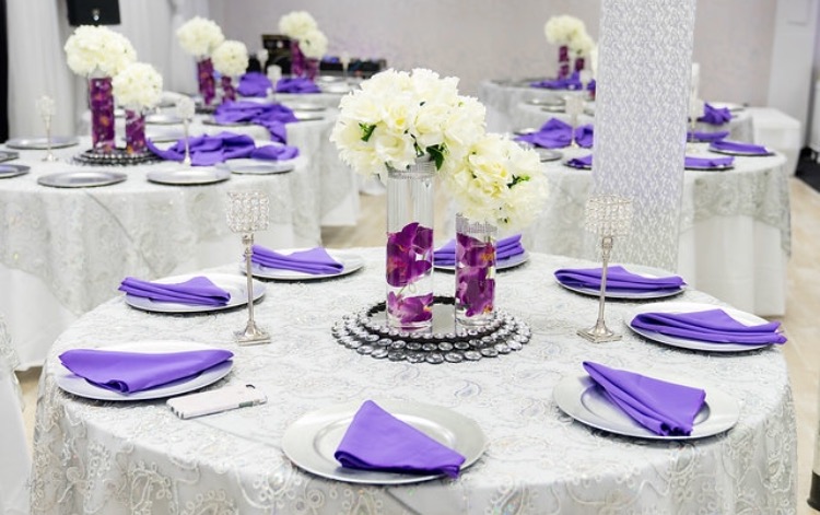 Elegant wedding reception table setting featuring white tablecloths, purple napkins, and centerpieces with white flowers and submerged purple flowers in glass vases at the Capitol Gallery.