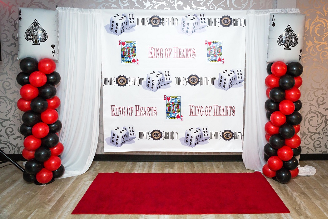 A party photo booth backdrop with a "king of hearts" theme, featuring playing card banners, dice decorations, and red and black balloon columns on each side. A red carpet leads to the backdrop set
