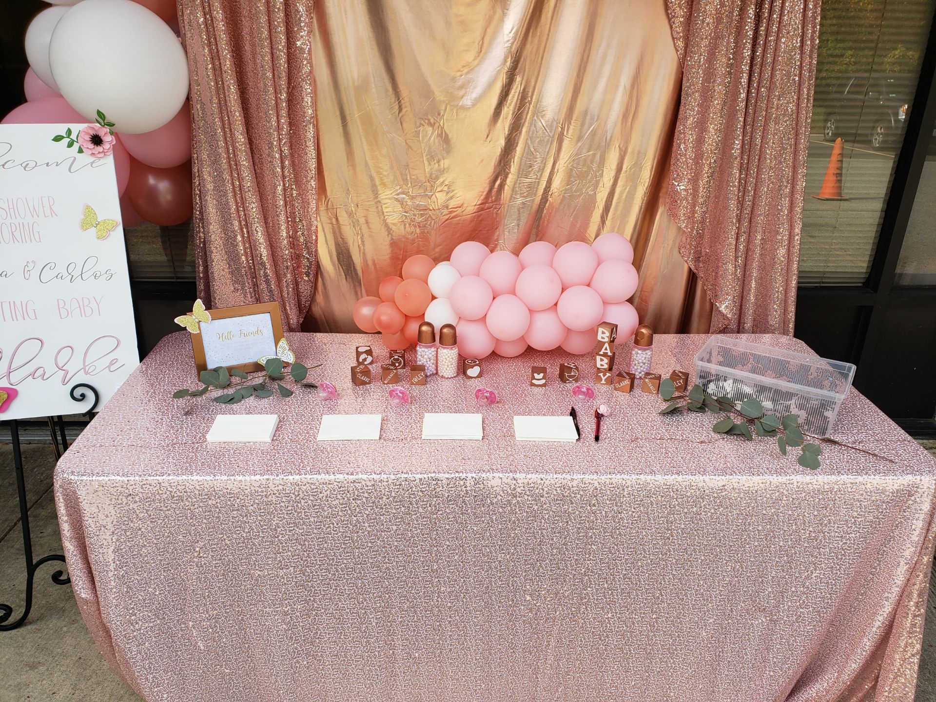 A table decorated in pink with a gold fabric backdrop, featuring a balloon arrangement and various small gifts and cards for a baby shower event at the Capitol Powerpack Gallery.