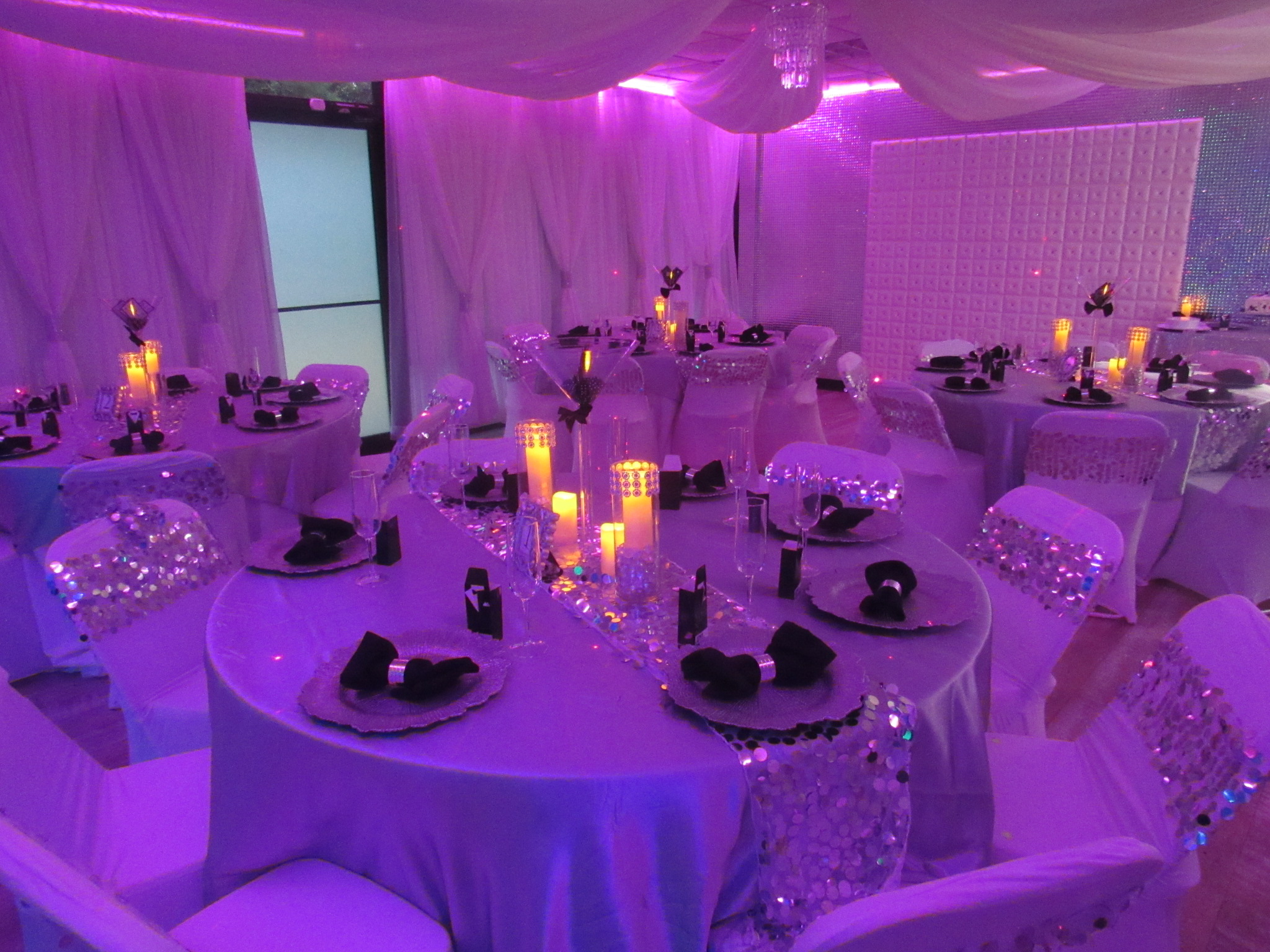 Elegant event space with round tables set for a formal dinner, featuring white tablecloths, black napkins, and glowing centerpieces, all under soft pink ambient lighting.