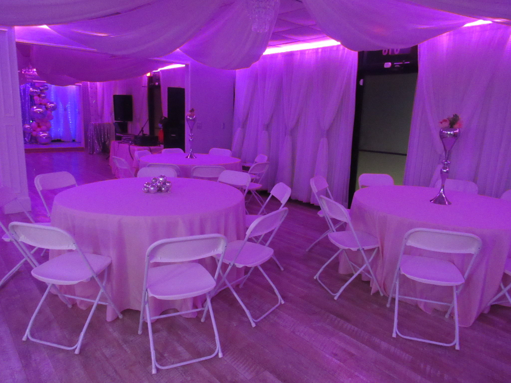 An event hall decorated with purple lighting featuring round tables draped with white tablecloths and surrounded by white chairs, awaiting guests.