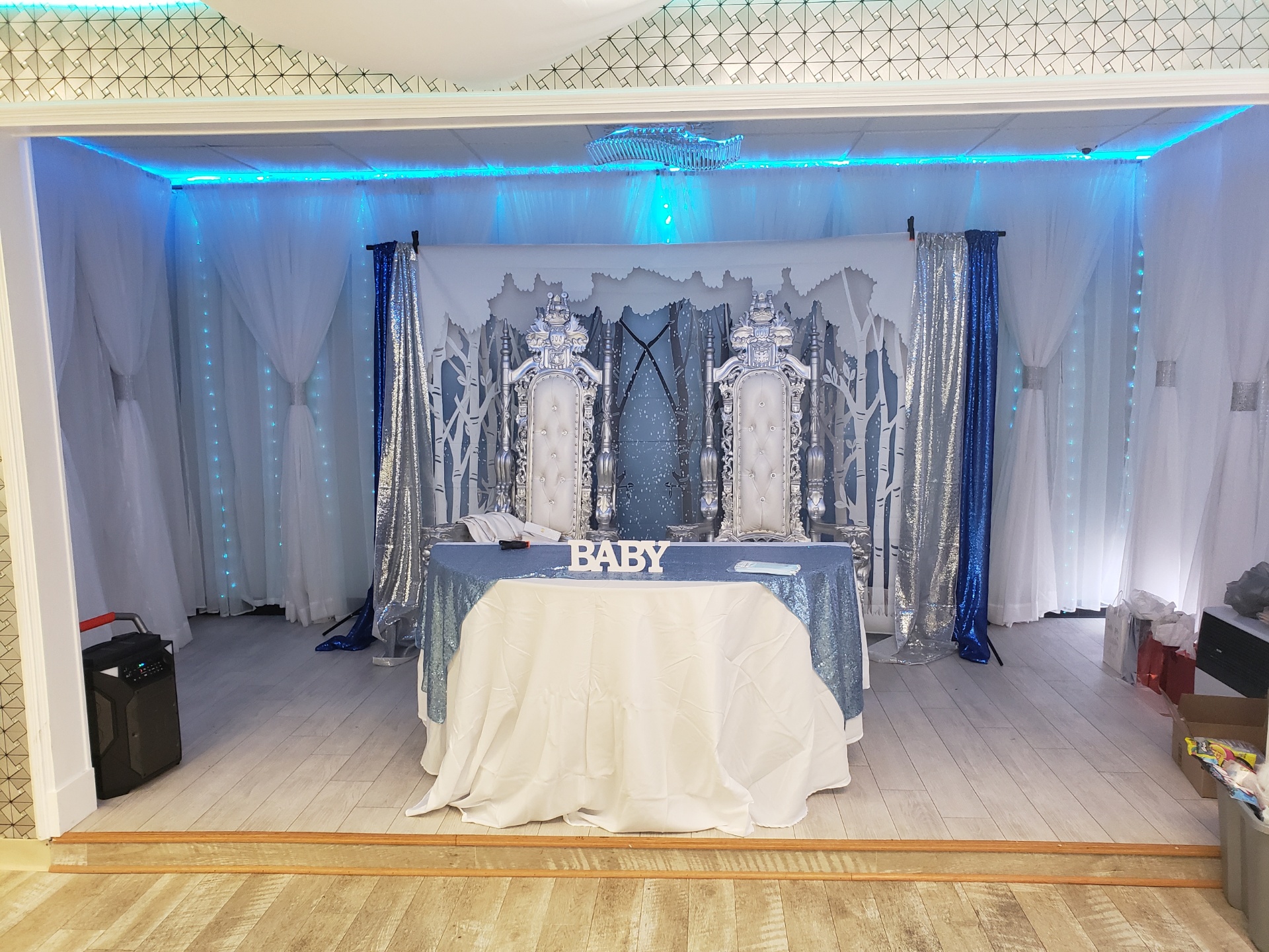 An elegantly decorated baby shower venue with two ornate chairs, a table draped in white, and "baby" spelled out in front of a blue-lit backdrop with white and blue curtains.