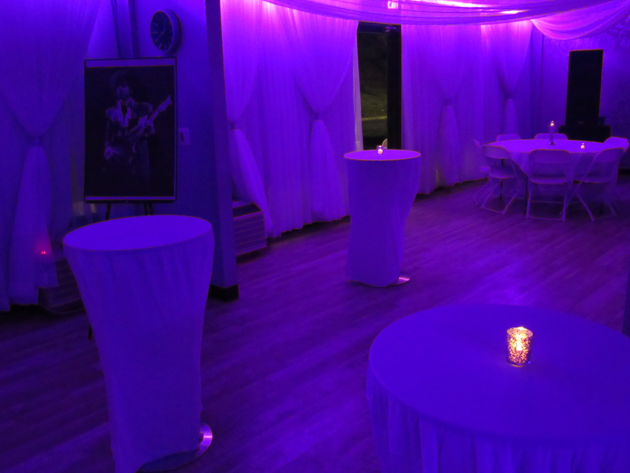 An event room illuminated in purple light with draped white curtains, several round tables covered with white cloths, and a framed picture of a guitar player.