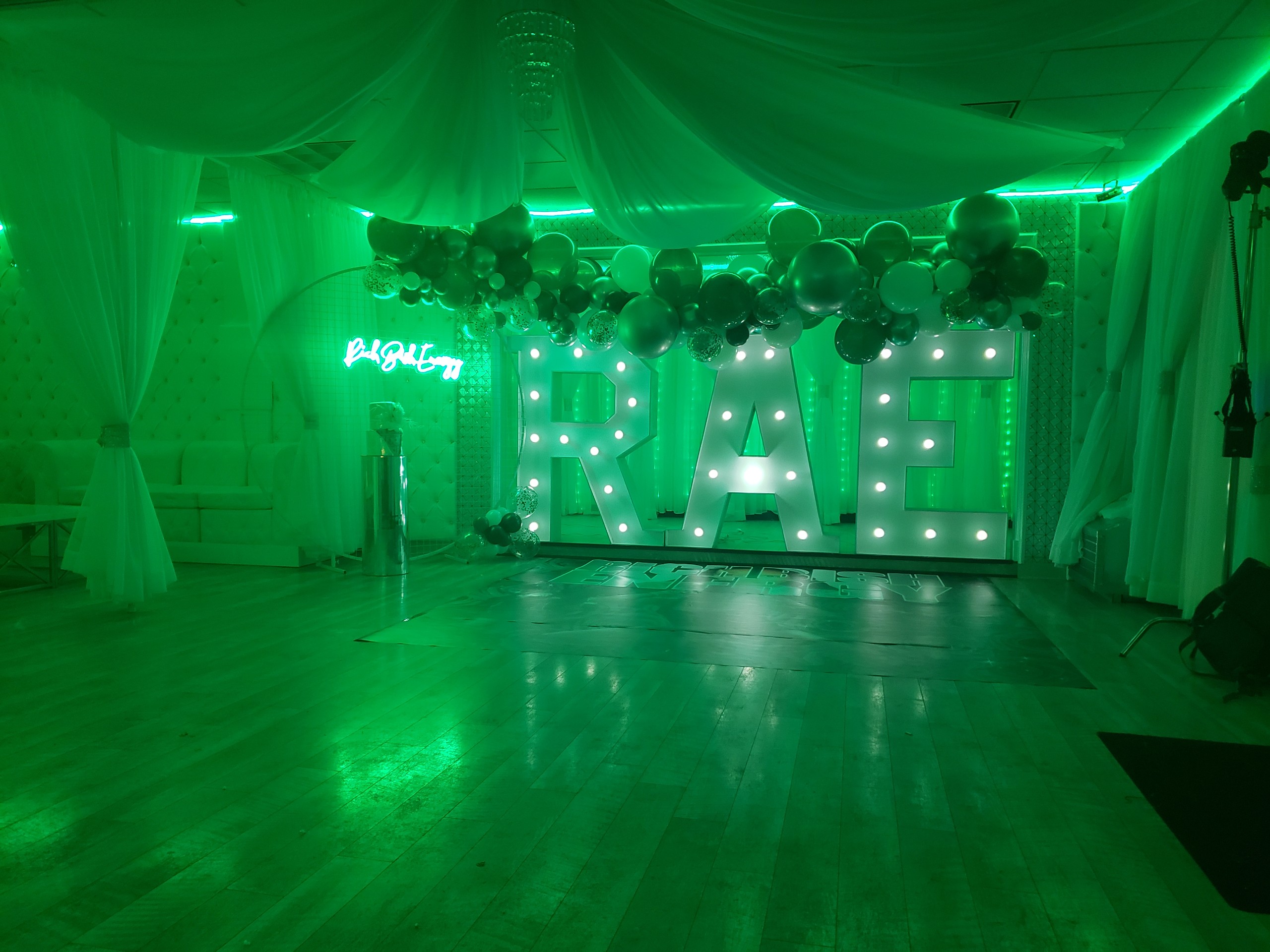 Decorated event hall with large illuminated letters spelling "rae" surrounded by black and white balloons, green lighting, and a neon sign.