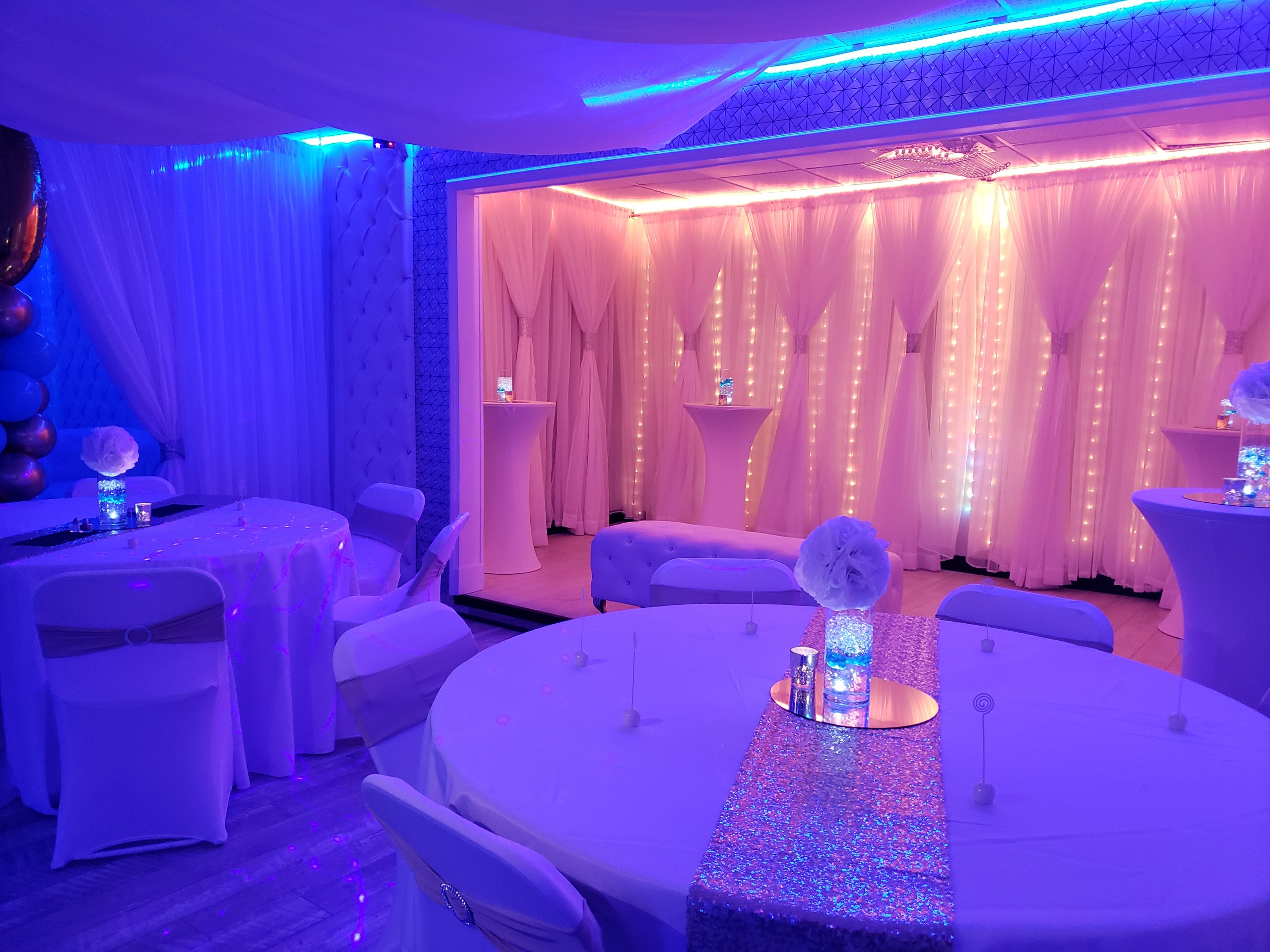 An elegant event hall with dim lighting decorated with blue and pink lights, white draped curtains, round tables with white covers, and a glittery centerpiece on each table.