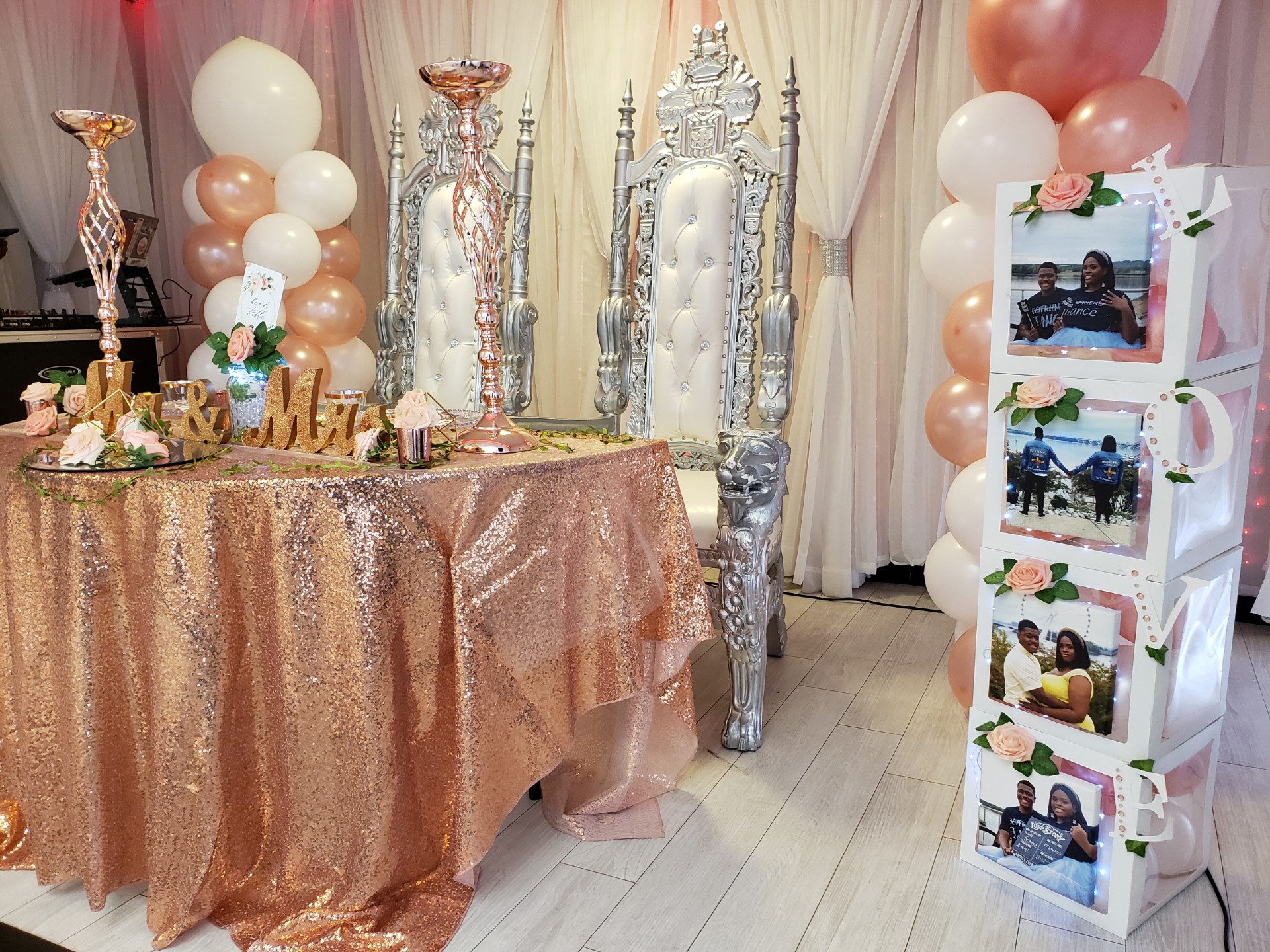 Elegant party setup featuring a table with a glittery rose gold cloth, adorned with candles, flowers, and gold decor. Two ornate silver columns and a Powerpack Gallery photo stand with balloons to