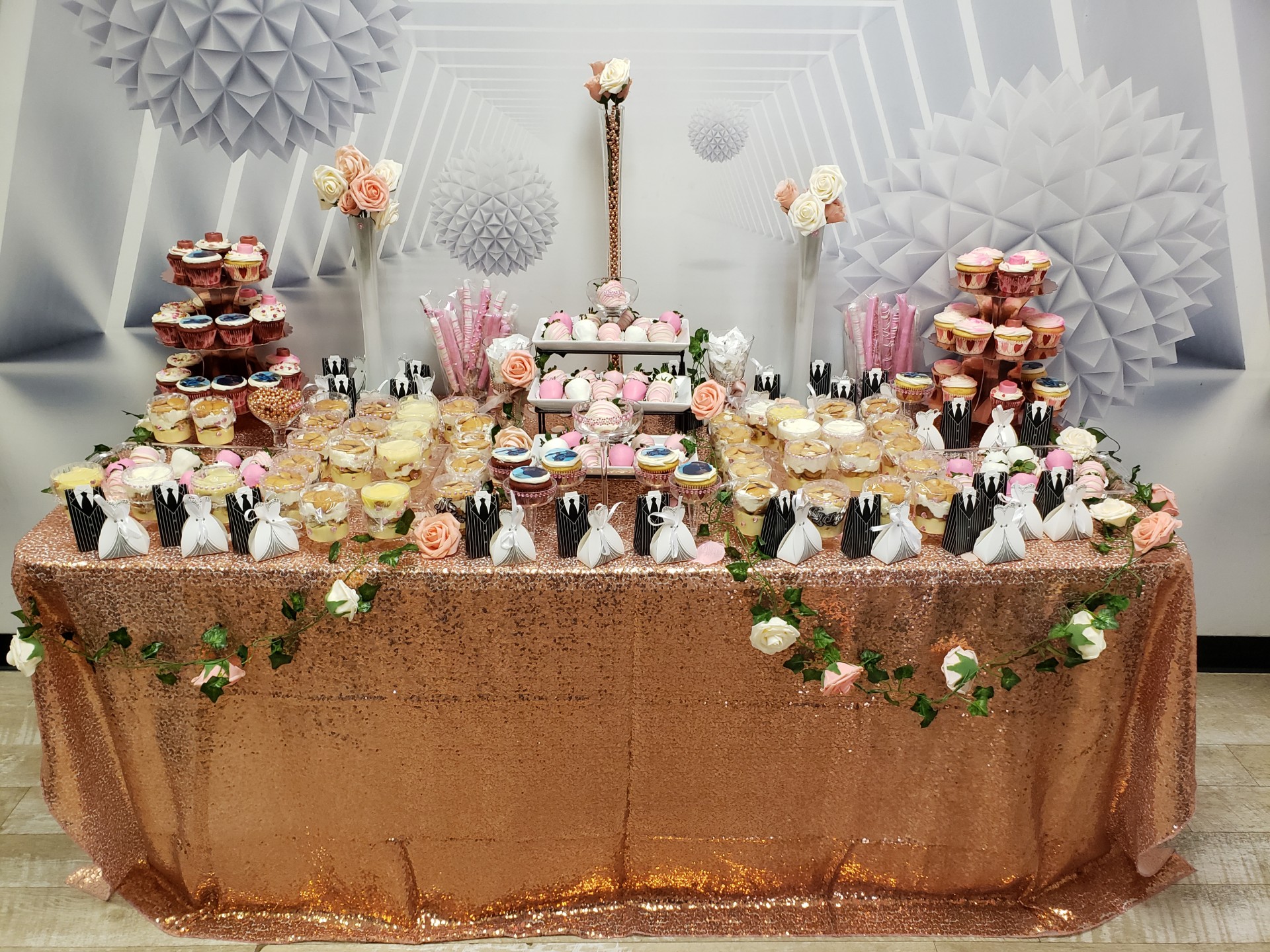 A beautifully arranged dessert table covered with a shimmering gold tablecloth, decorated with cupcakes, macarons, and other sweets, adorned with floral accents and elegant decor in a gallery setting.