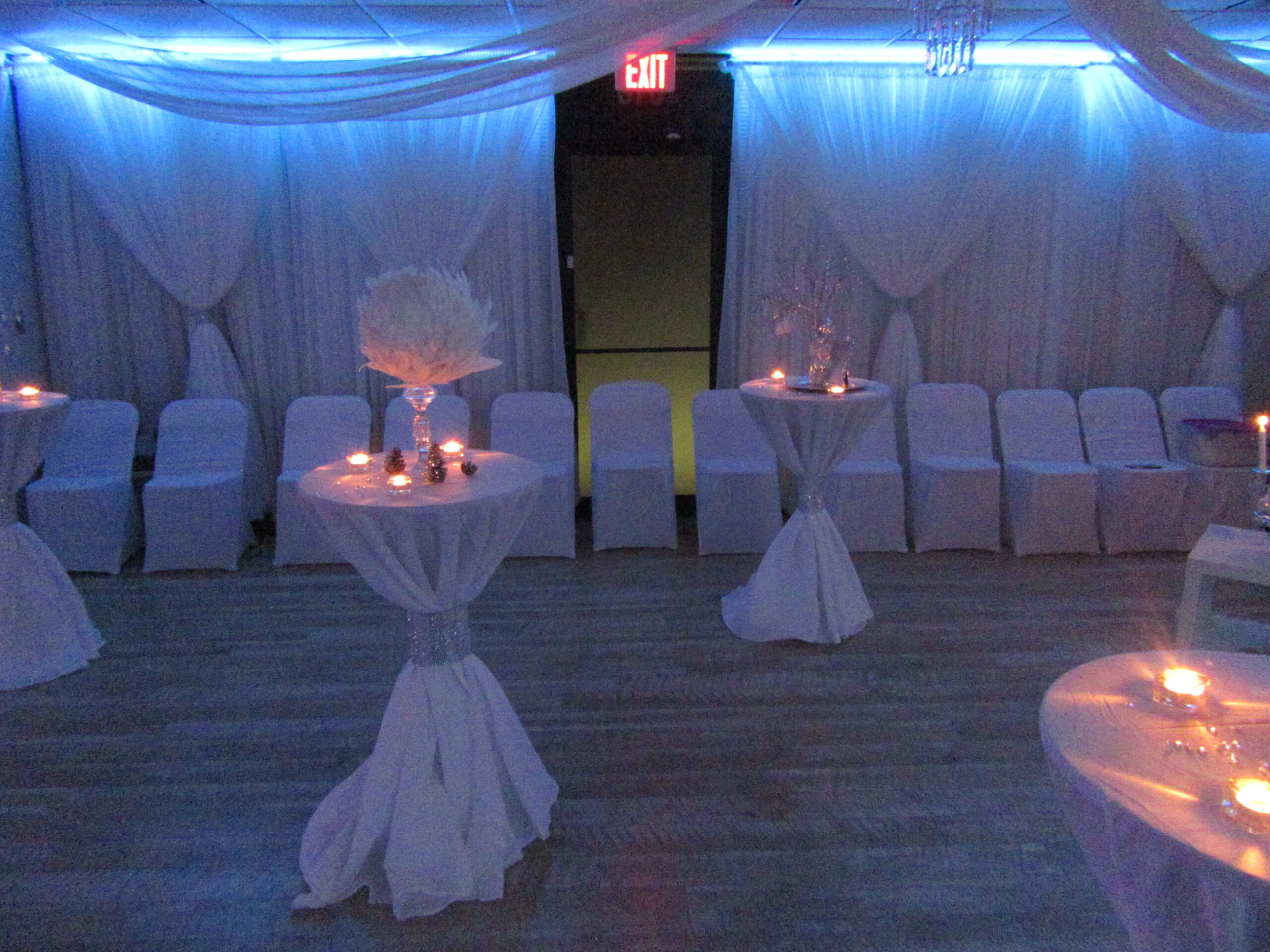 A dimly lit event hall decorated with white drapes and chair covers, featuring round tables with white tablecloths, centerpieces with glowing candles, and one yellow chair in the background.