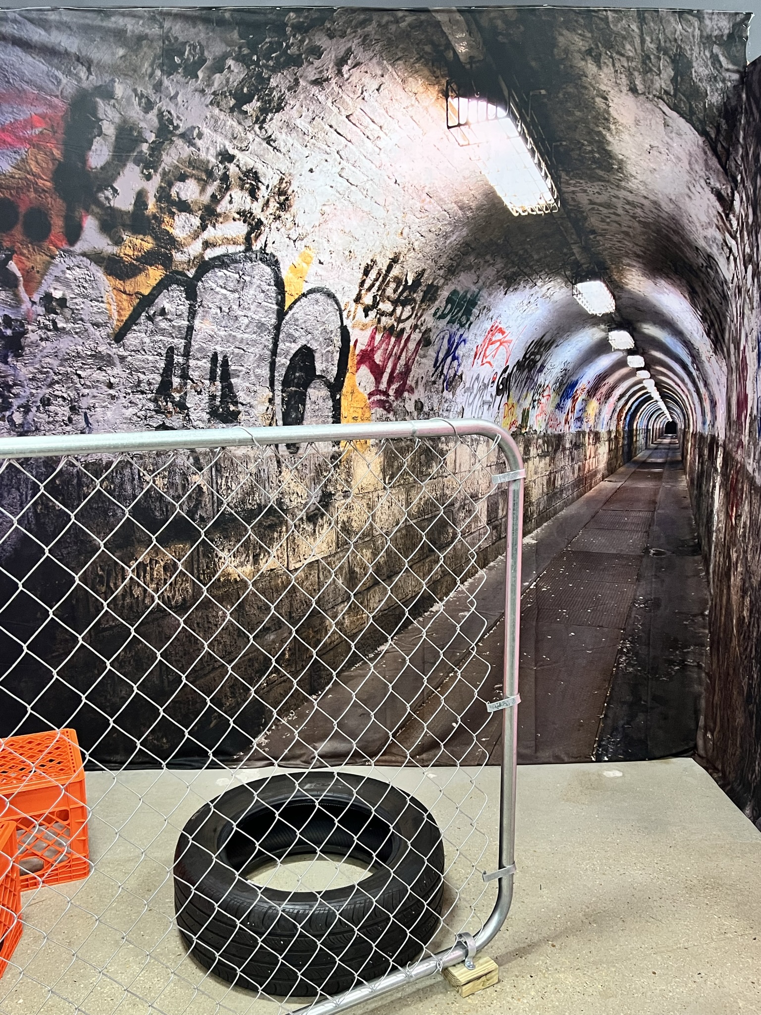 A long urban tunnel featuring graffiti on its arched walls, illuminated intermittently by overhead lights, partially obstructed at the entrance by a metal barricade and a tire.