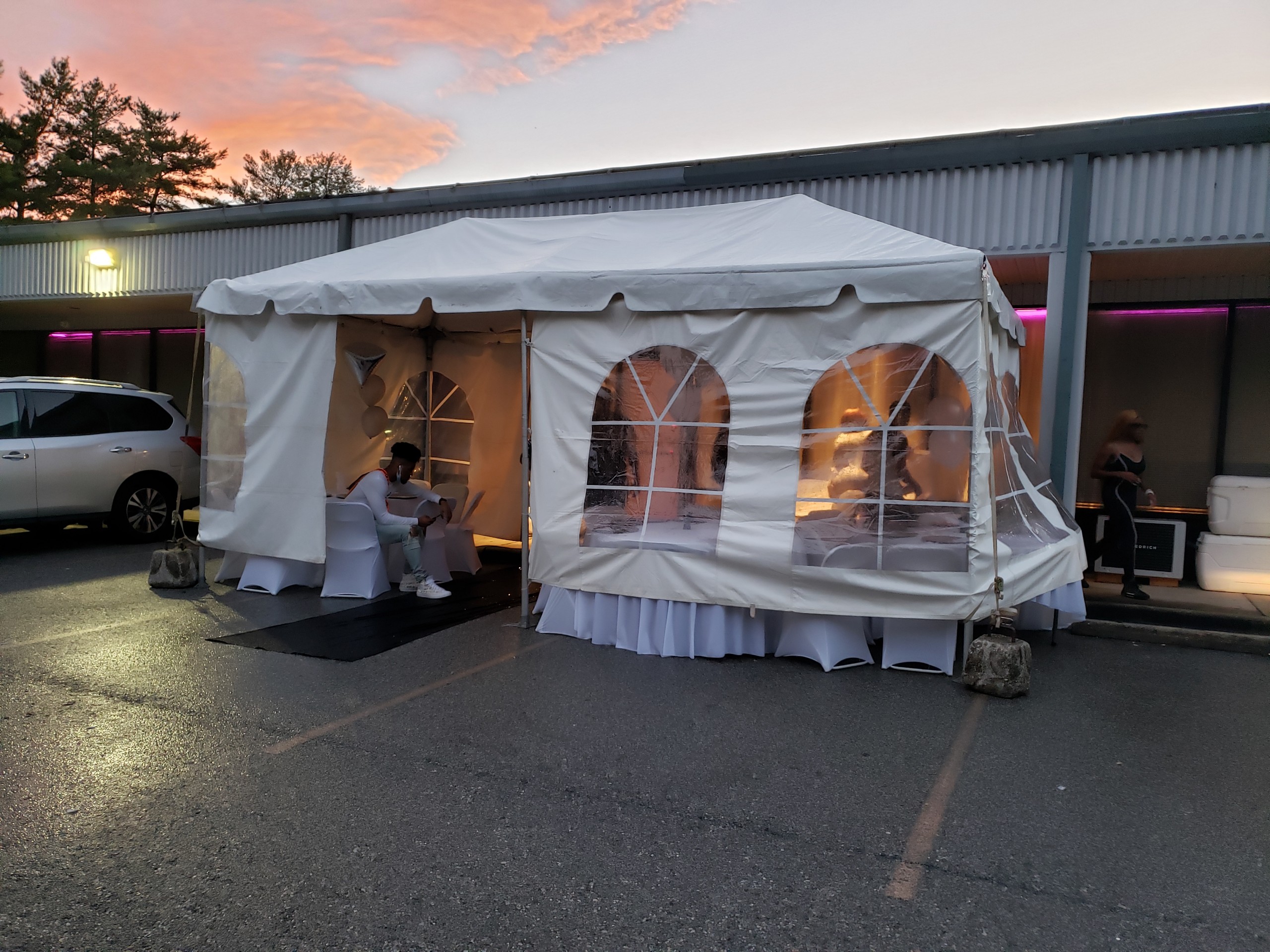 A white outdoor tent with transparent window panels set up in the parking lot of the Capitol Powerpack Gallery, illuminated within, as people dine inside during a colorful sunset.