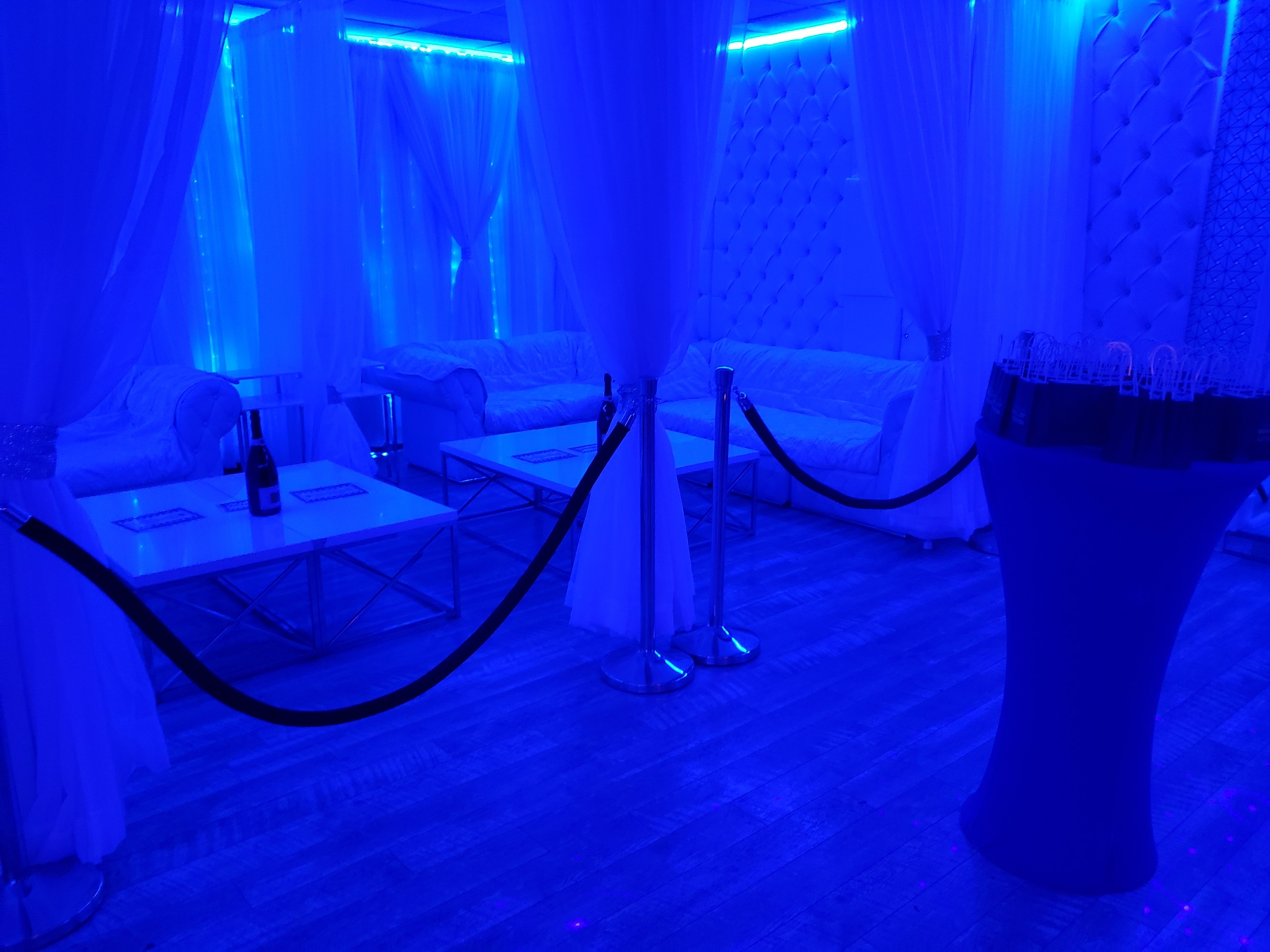 A blue-lit lounge area with draped white curtains, plush sofas, a glass table with champagne, and a nearby standing cocktail table under soft blue lighting.