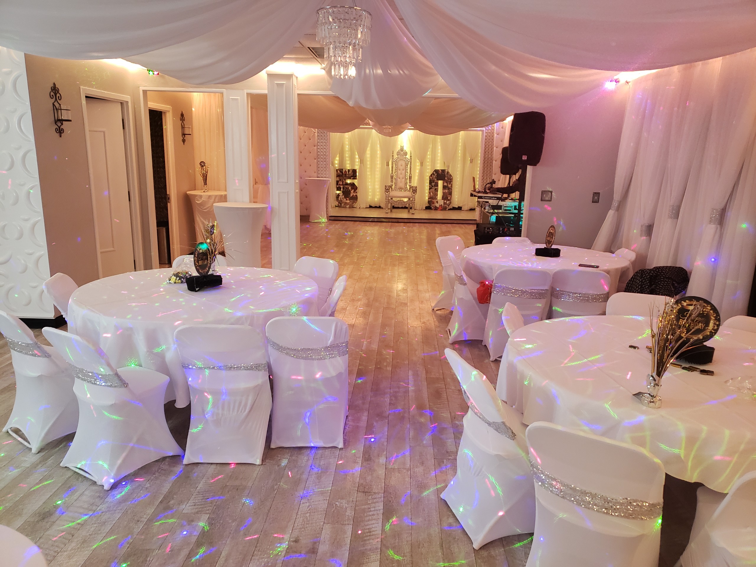 Elegant banquet hall decorated for an event with white draped tables and chairs; multi-colored lights project on floor, large chandeliers overhead, and mirrored walls enhance spacious interior.