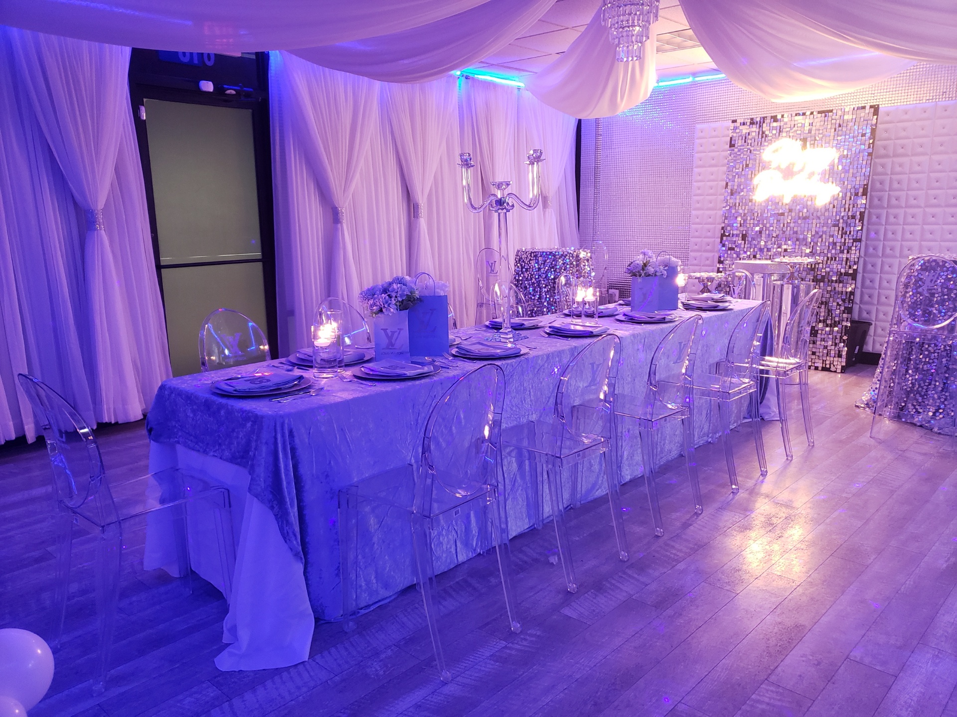 Elegant event setting with a long table adorned in blue tablecloth, clear chairs, and elaborate place settings, under purple lighting, with draped white curtains and decorative panels in the background.