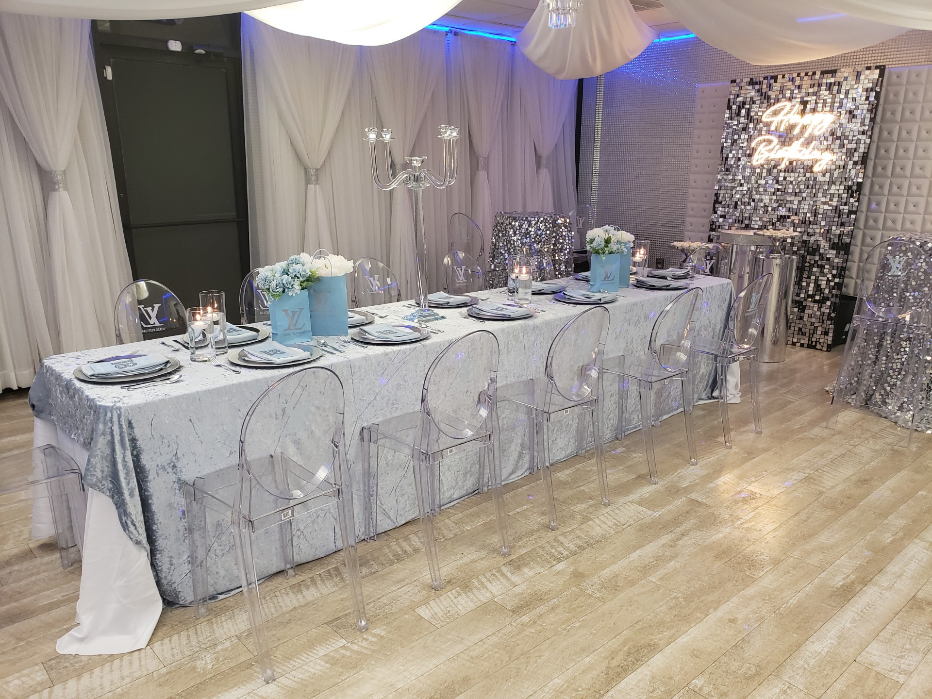 Elegant event table setting featuring transparent chairs, blue-themed decor with floral centerpieces, and a lit-up 'happy birthday' backdrop in a room with silver curtains.