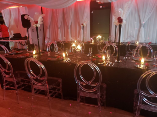 Elegant event setup featuring a long table with sparkling settings, transparent chairs, and dramatic red lighting. White curtains and feather decorations add a luxurious touch to the homepage.