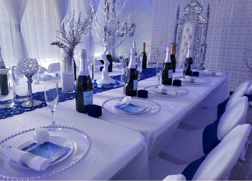 Elegant dining setup with a blue and white theme, featuring a long table adorned with champagne bottles, crystal glasses, and decorative silver elements under soft lighting.