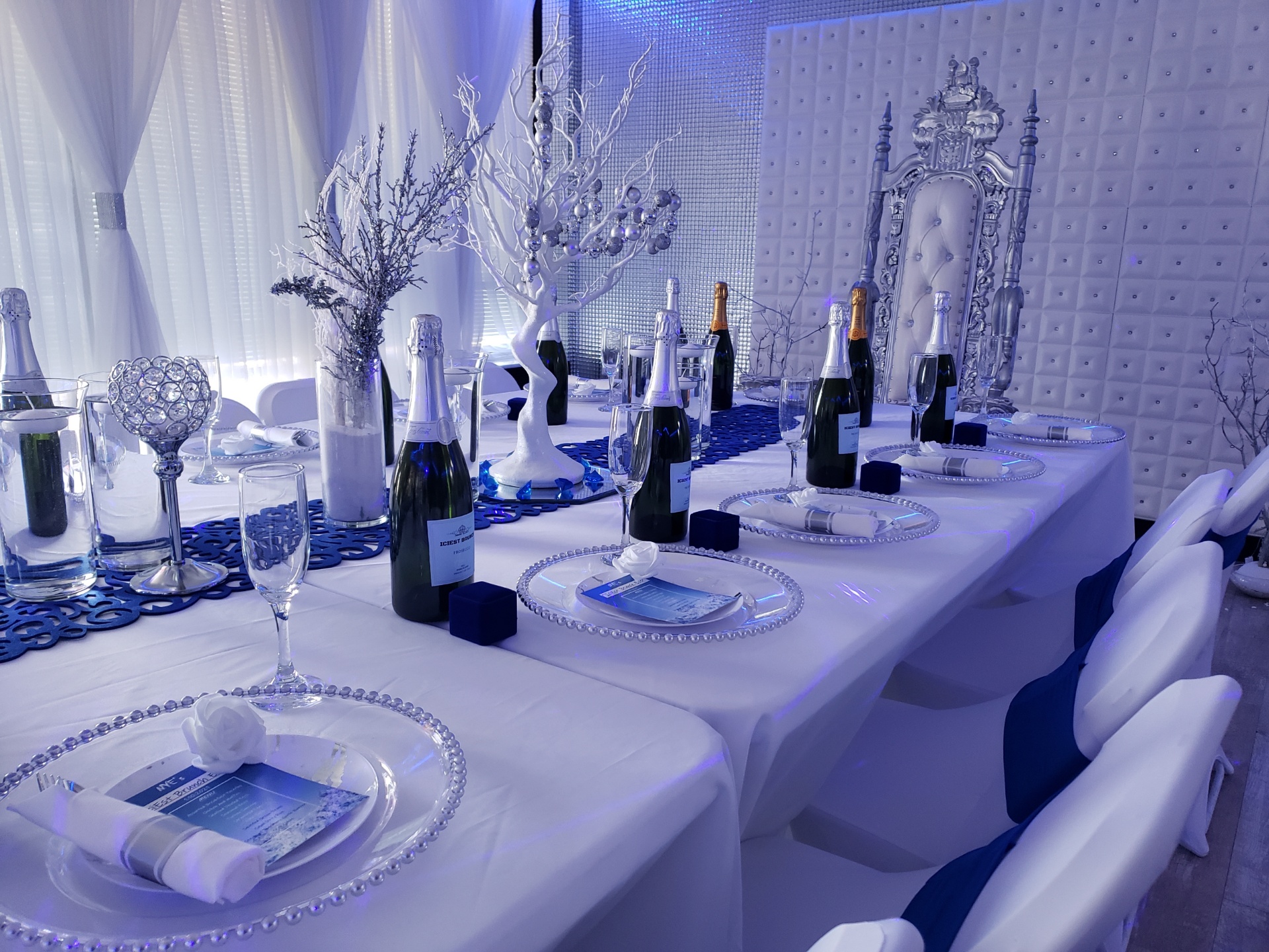 Elegant banquet table setup with white and silver decor, featuring champagne bottles, crystal glassware, and ornamental trees, in a room with blue lighting.