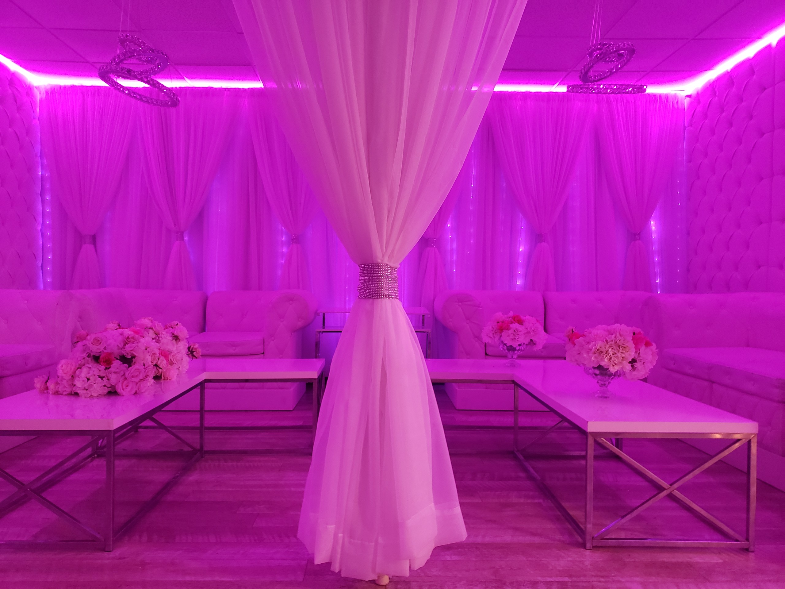 An elegant event space with pink lighting and white decor, featuring plush sofas, floral arrangements, draped curtains, and crystal chandeliers.