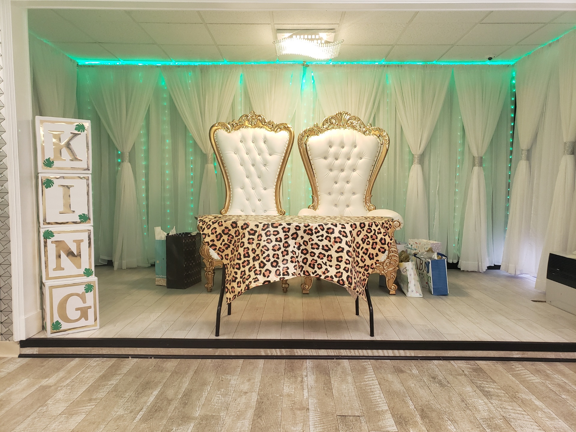 Two ornate white and gold chairs set on a stage with a leopard print cushion, surrounded by white drapes and mint green lighting. a sign reading "king" stands to the left.