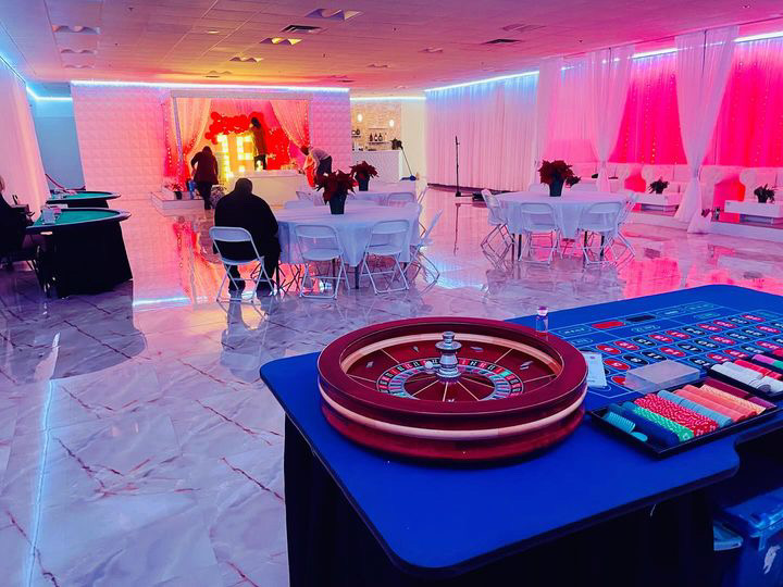 An indoor party venue with a casino theme, featuring a large roulette table in the foreground, round dining tables, and colorful lighting highlighting a stage area in the back.