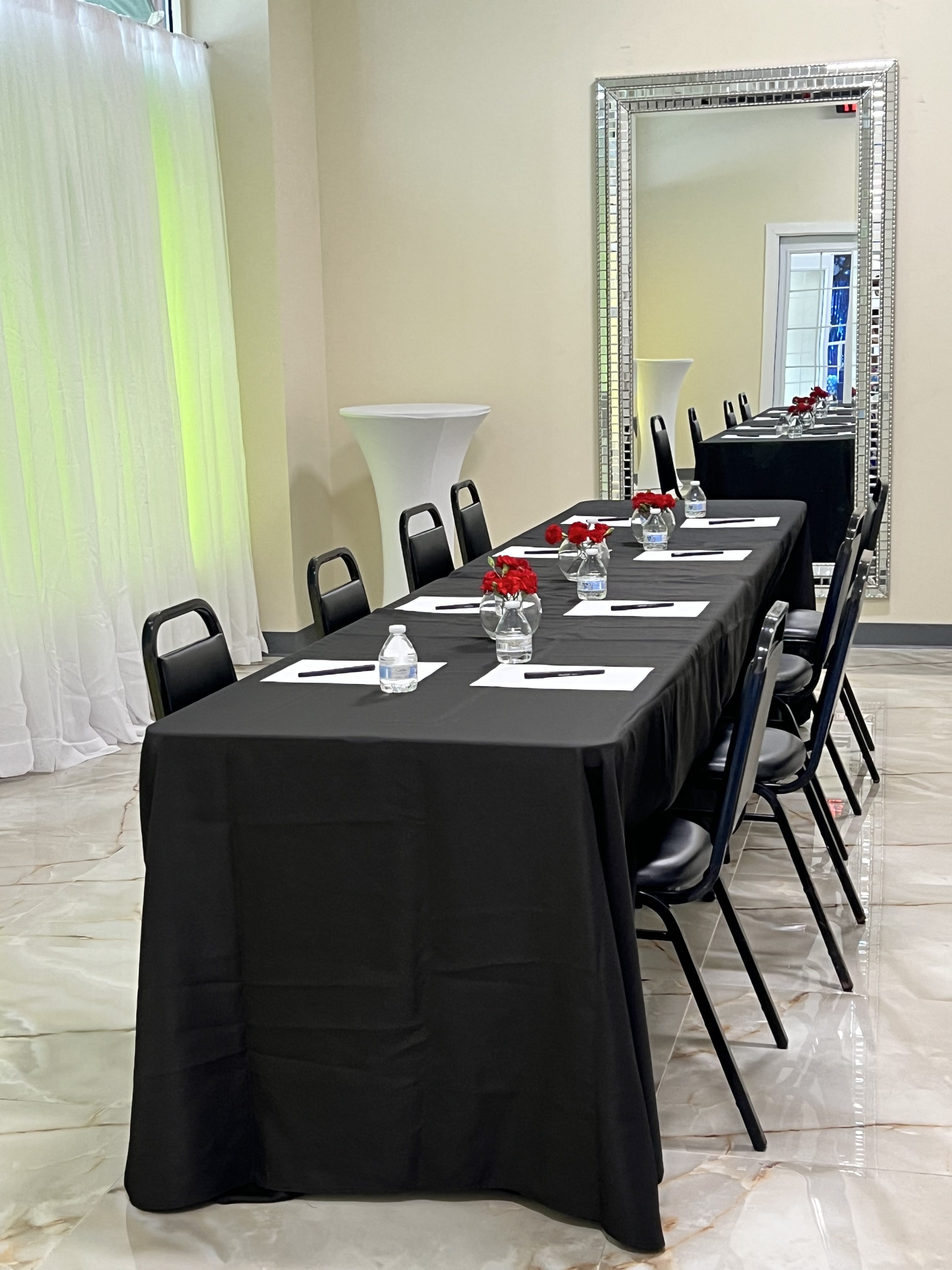 A banquet hall setup with a long table covered in a black tablecloth, featuring neatly arranged black chairs, white plates, water bottles, and small red flower centerpieces.