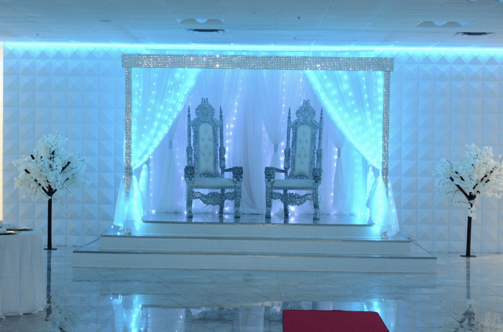Elaborate wedding stage with two ornate chairs, illuminated by blue lights and white drapes, flanked by decorative white trees in a grand hall.