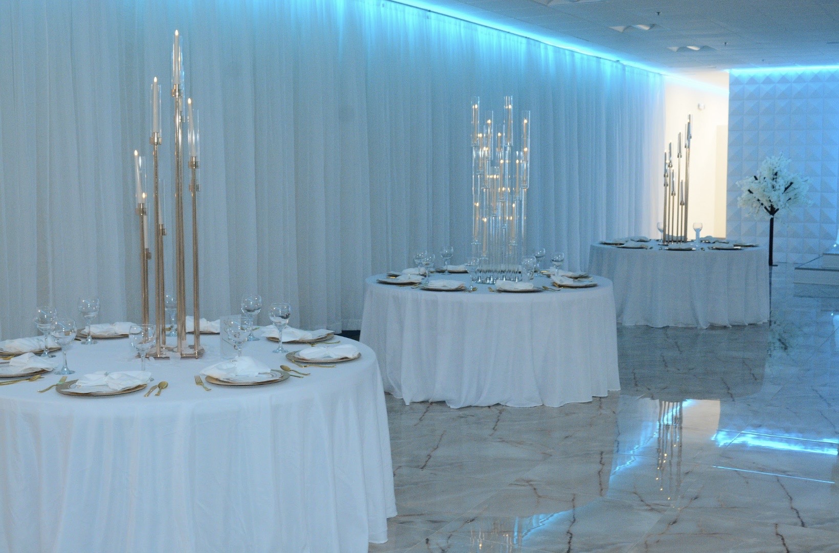 Elegant dining setup featuring tables with white tablecloths, gold-rimmed plates, and crystal glassware, complemented by tall candle stands and ambient blue lighting.