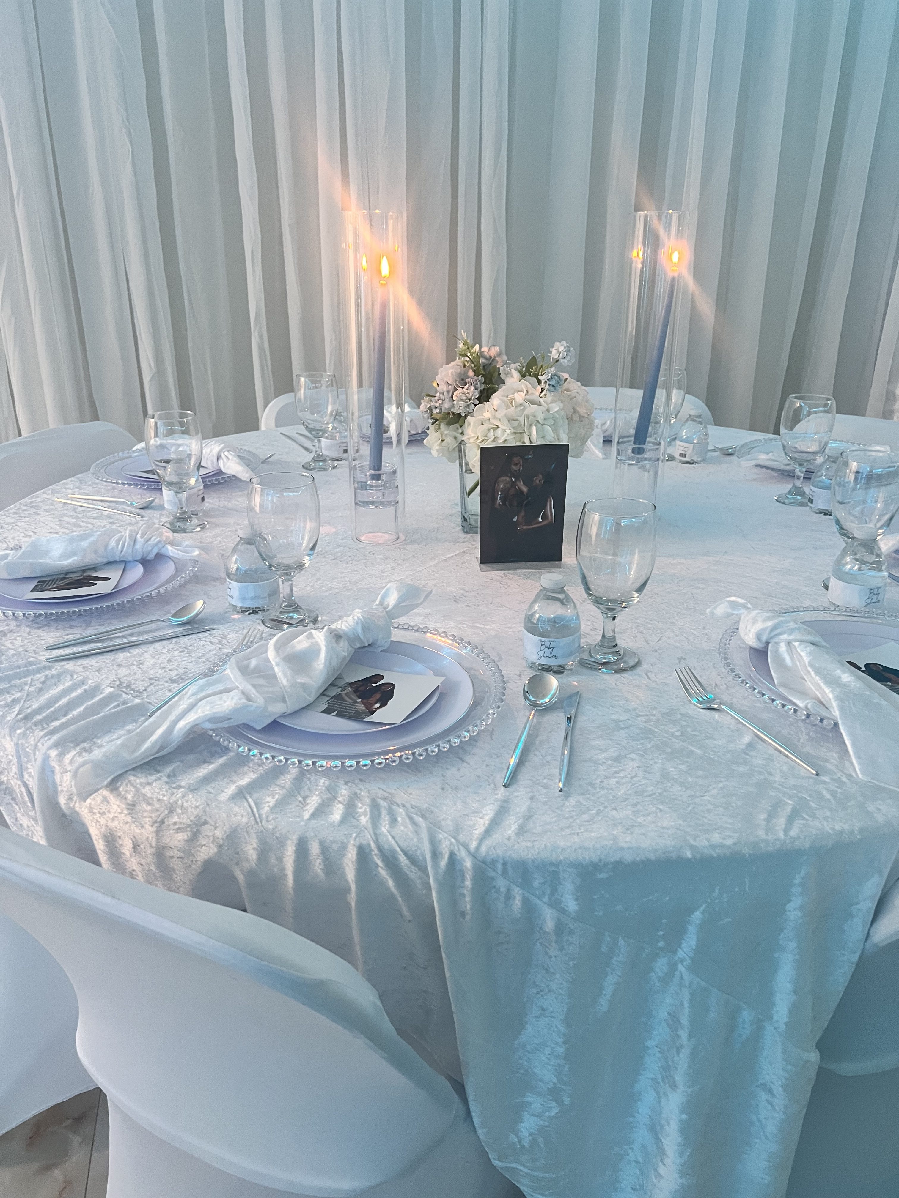 Elegant dining setup for a wedding with a white theme. table features floral arrangements, candles, set plates, and glasses, set against draped white curtains.