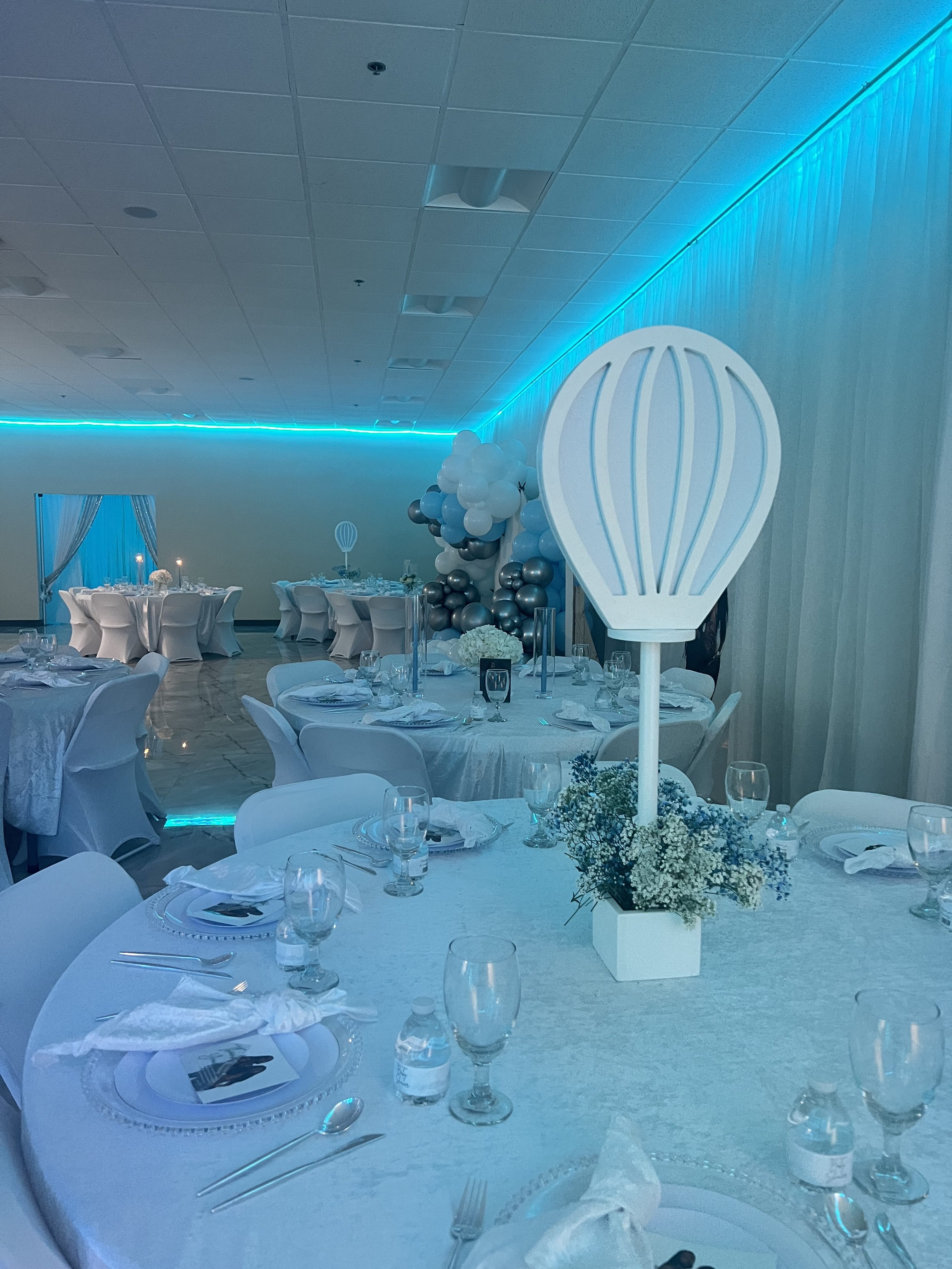 An elegant event setup in a room with blue lighting, featuring round tables dressed in white with centerpieces, glassware, and themed balloon decorations.