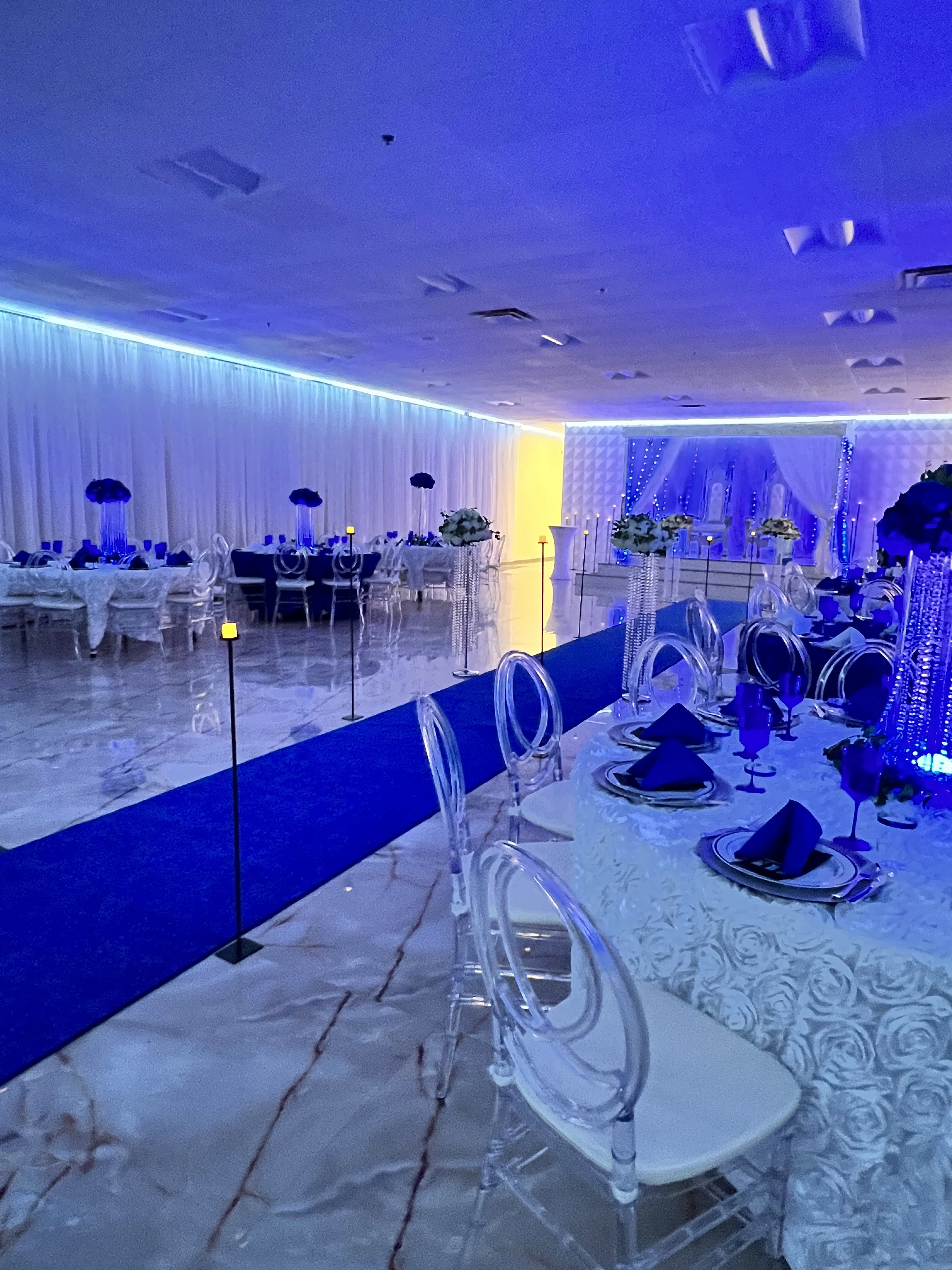 A banquet hall illuminated in blue light, featuring elegant round tables set with navy napkins and clear chairs on a glossy marble floor, leading to a yellow-lit stage at the far end.