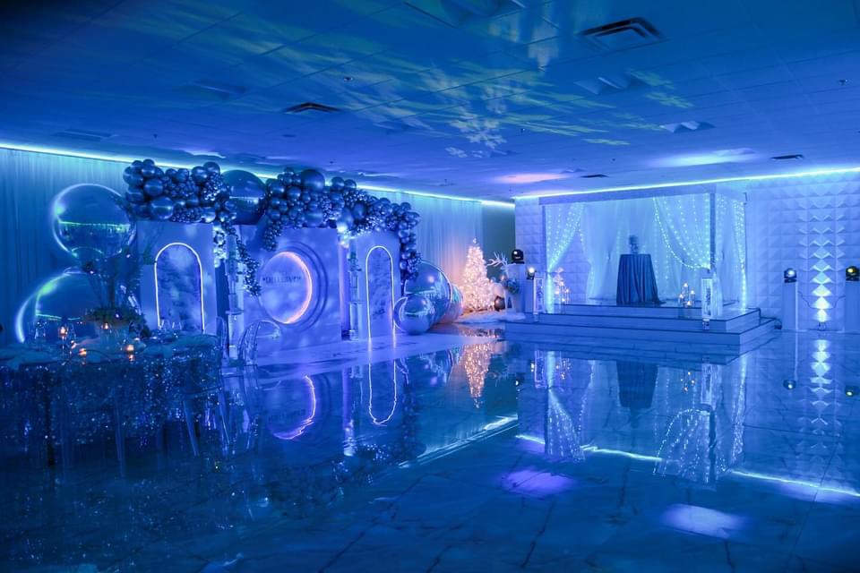 A luxurious event hall illuminated by blue lights, featuring reflective floors, elegant balloon decorations, and a stage with curtains.
