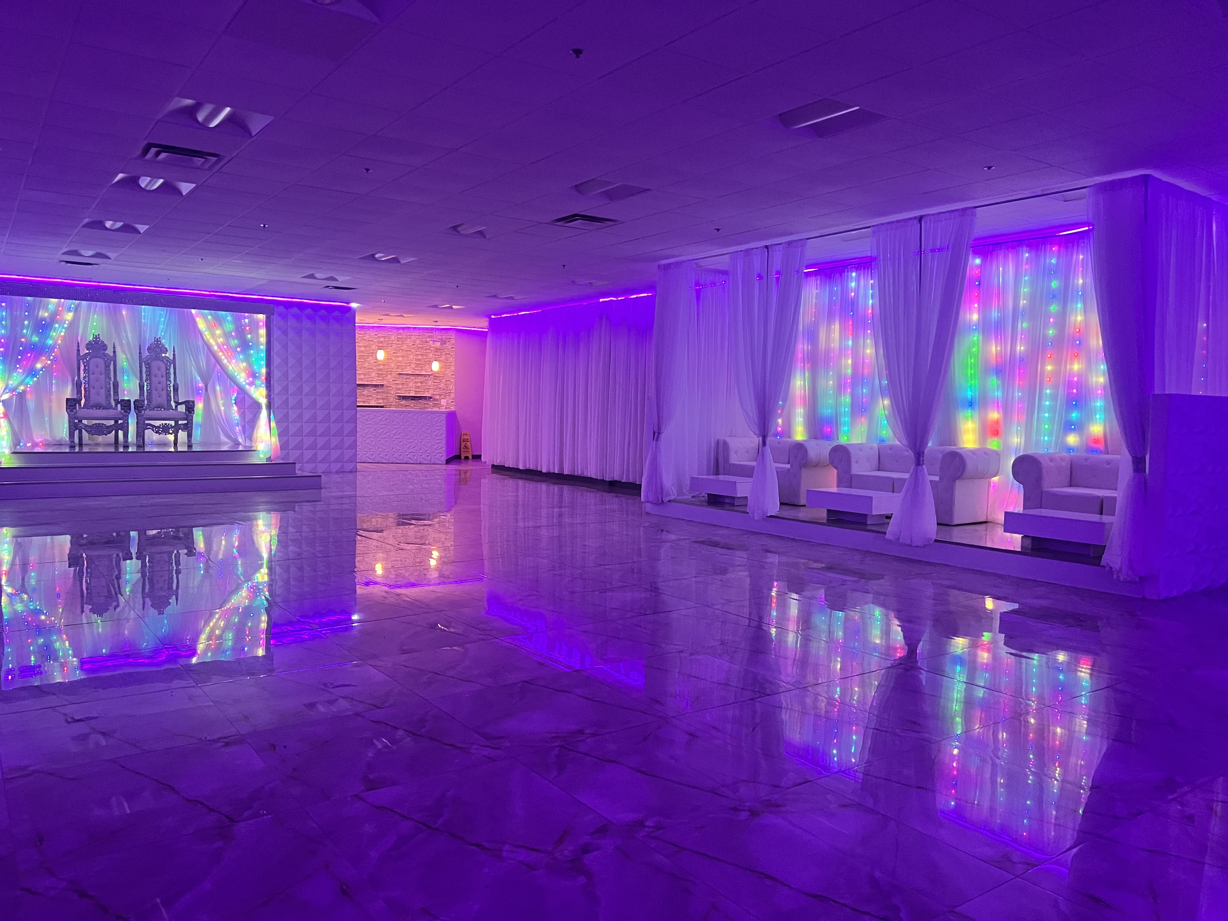 An event hall illuminated with purple and pink lights, featuring elegant white drapes and furnishings, with a glossy reflective floor enhancing the vibrant ambiance.