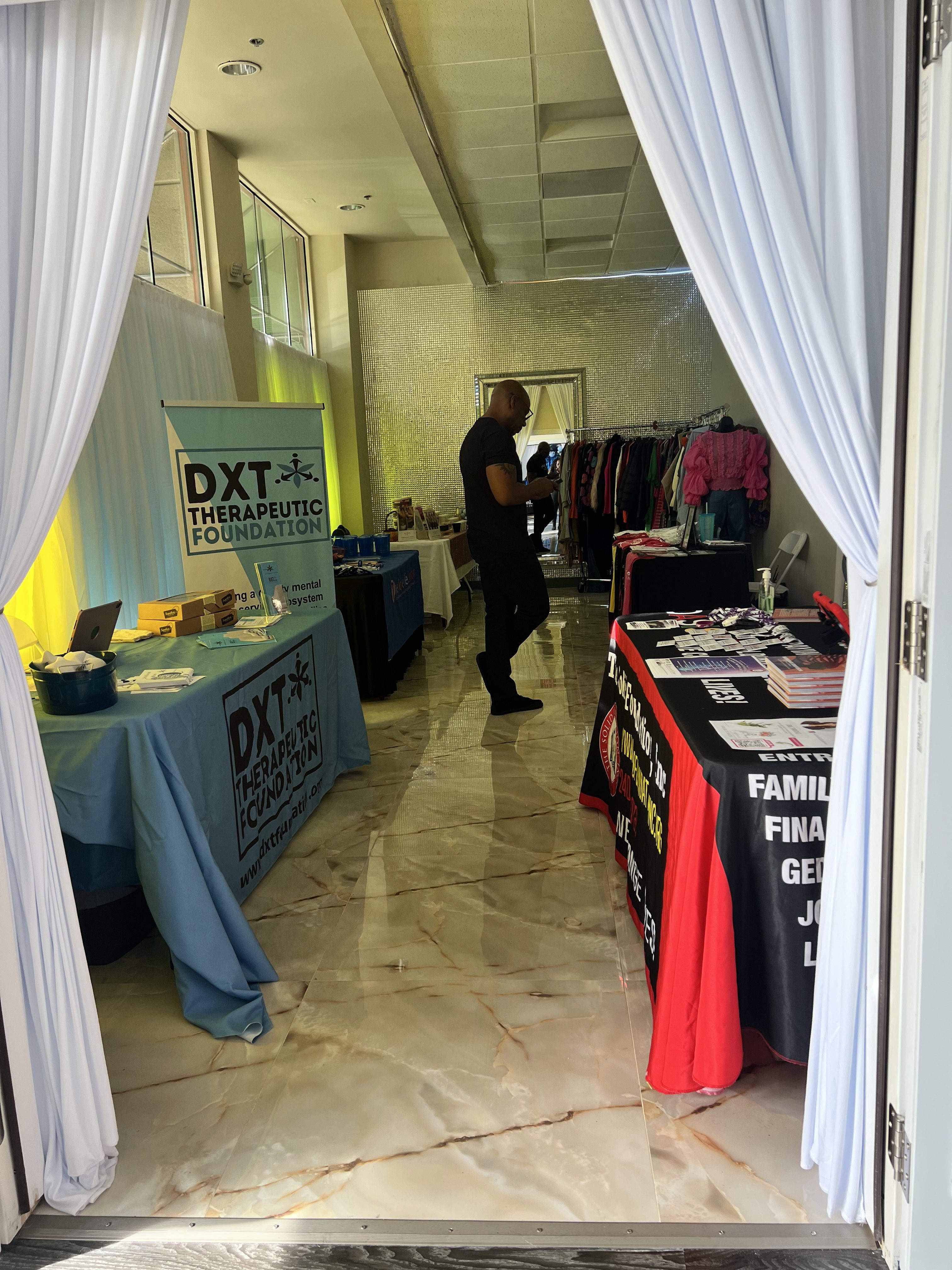 A man stands near a series of informational booths inside a room with marble flooring, flanked by open white curtains. booths showcase colorful banners and various informational materials.