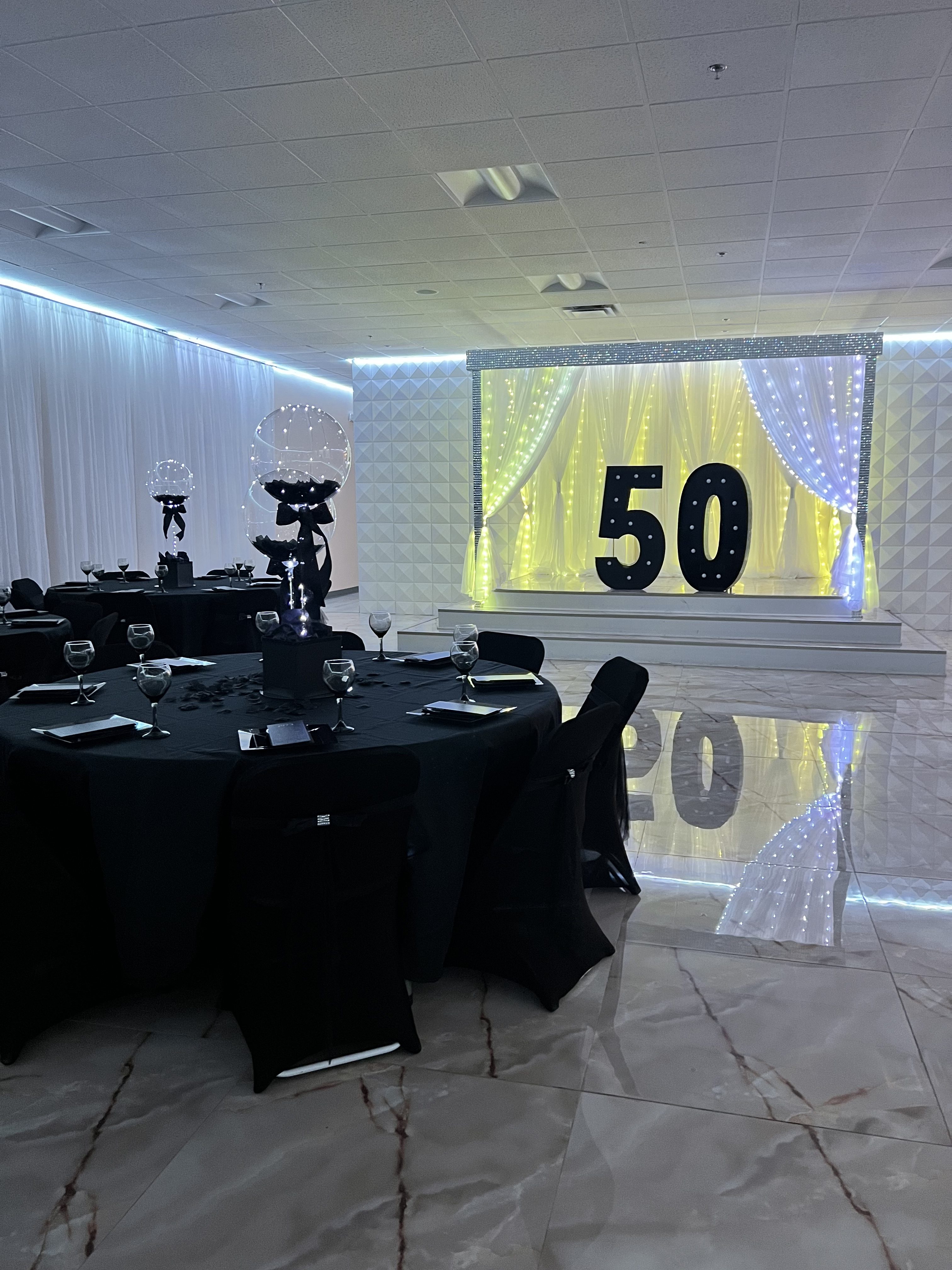 Elegant event space decorated for a 50th celebration with a large "50" lit up on the back wall, black tablecloths on round tables, reflective floor, and chic lighting.
