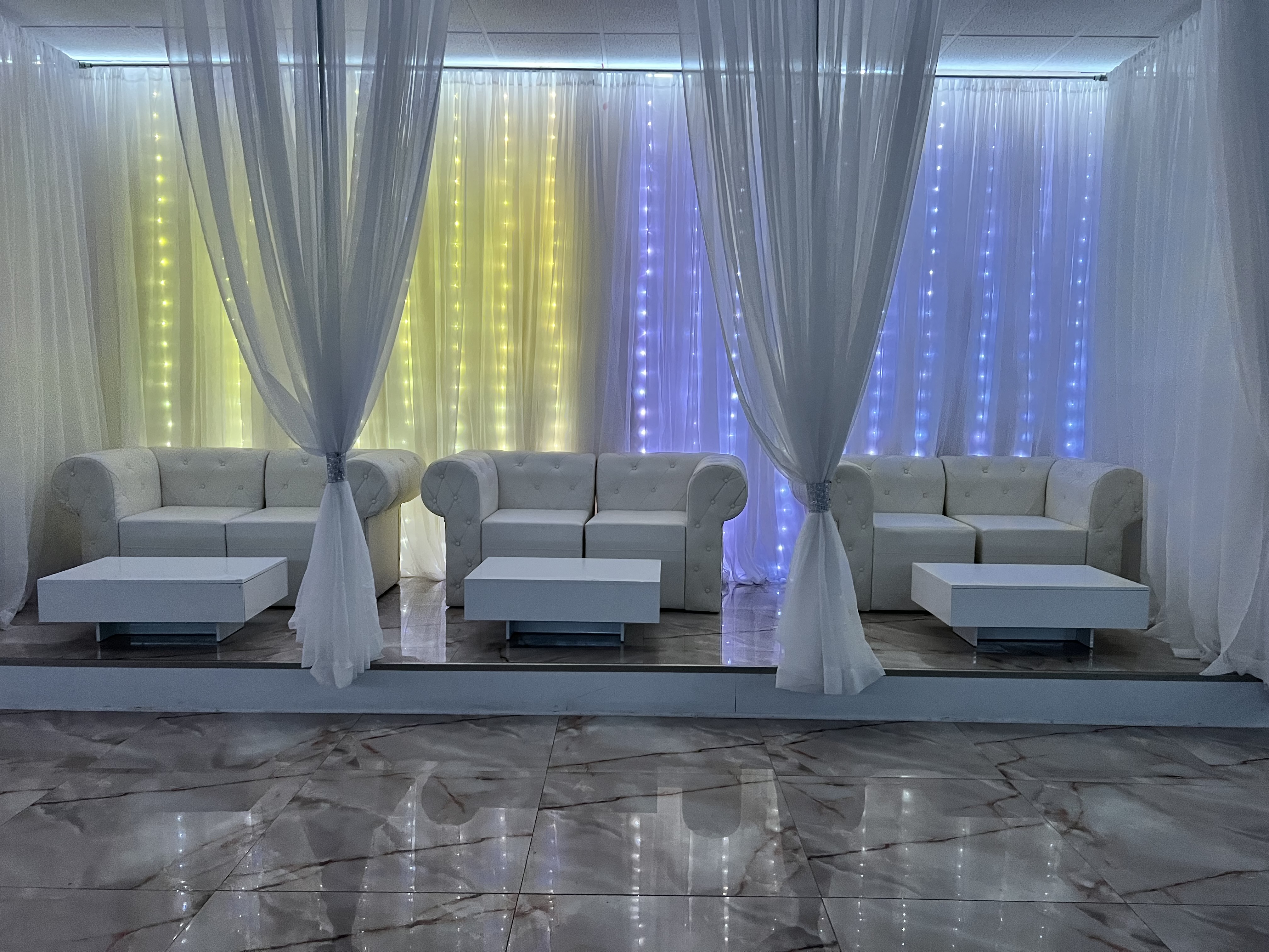 Event hall with white couches and small tables set against a wall with curtains and colorful vertical light strips behind. the floor is glossy and the lighting creates a festive ambiance.