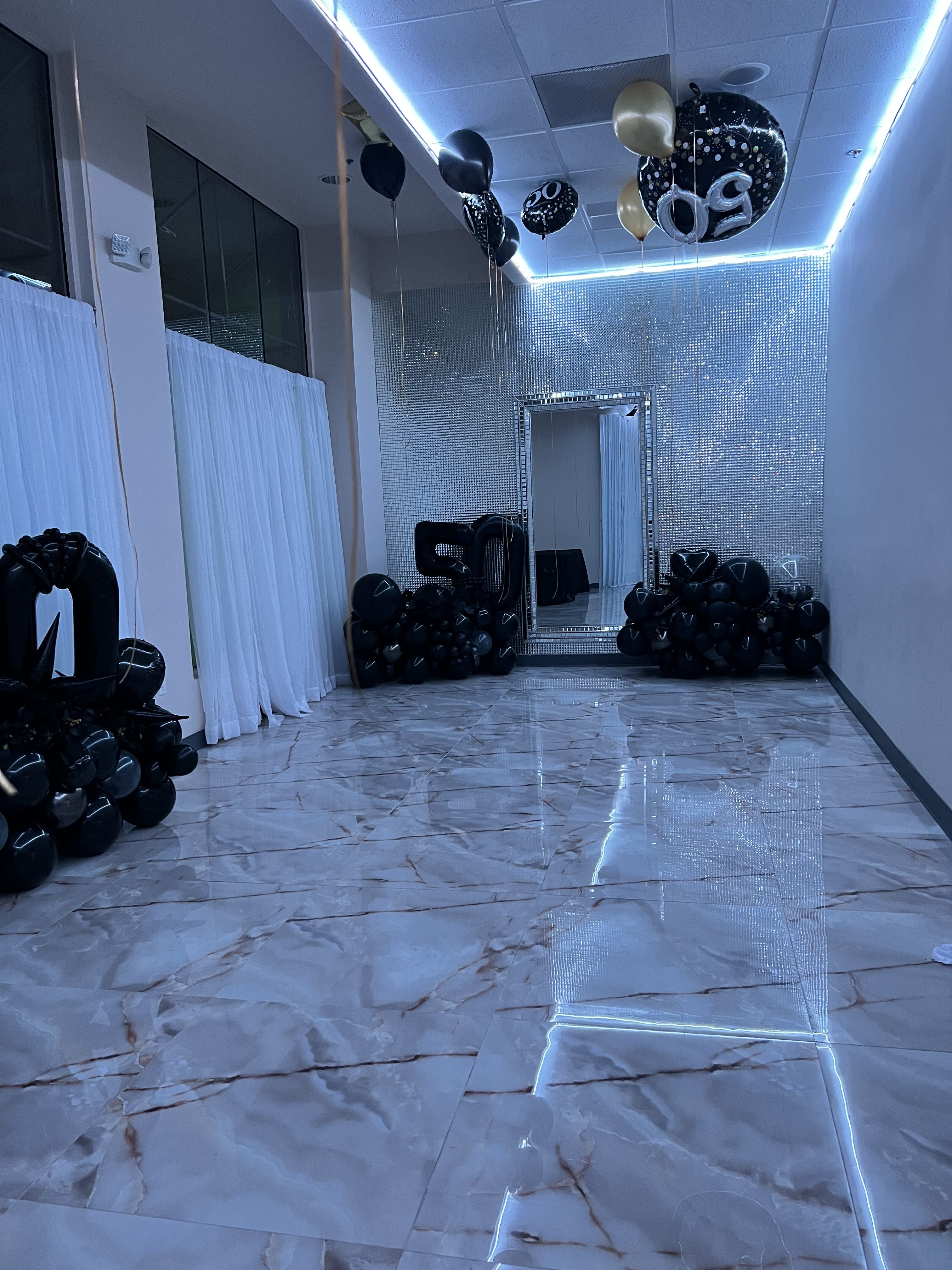 A decorated hallway with black and gold balloons, white curtains, and a lit-up ceiling leading to an open door. marble flooring adds to the luxe ambiance.