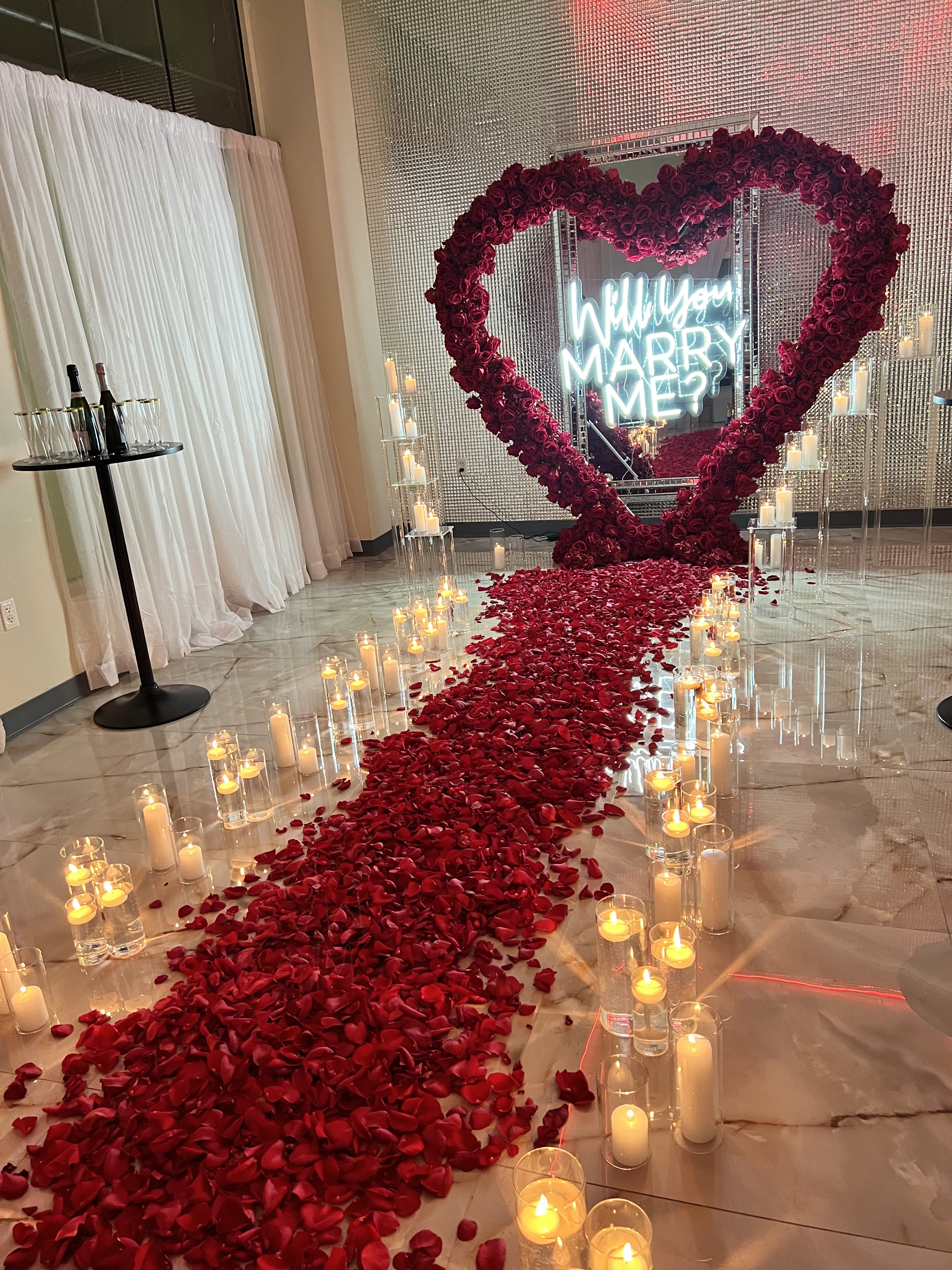 A romantic marriage proposal setup featuring a large heart-shaped frame decorated with rose petals, surrounded by lit candles, leading to a neon sign saying "will you marry me?" in cursive script.