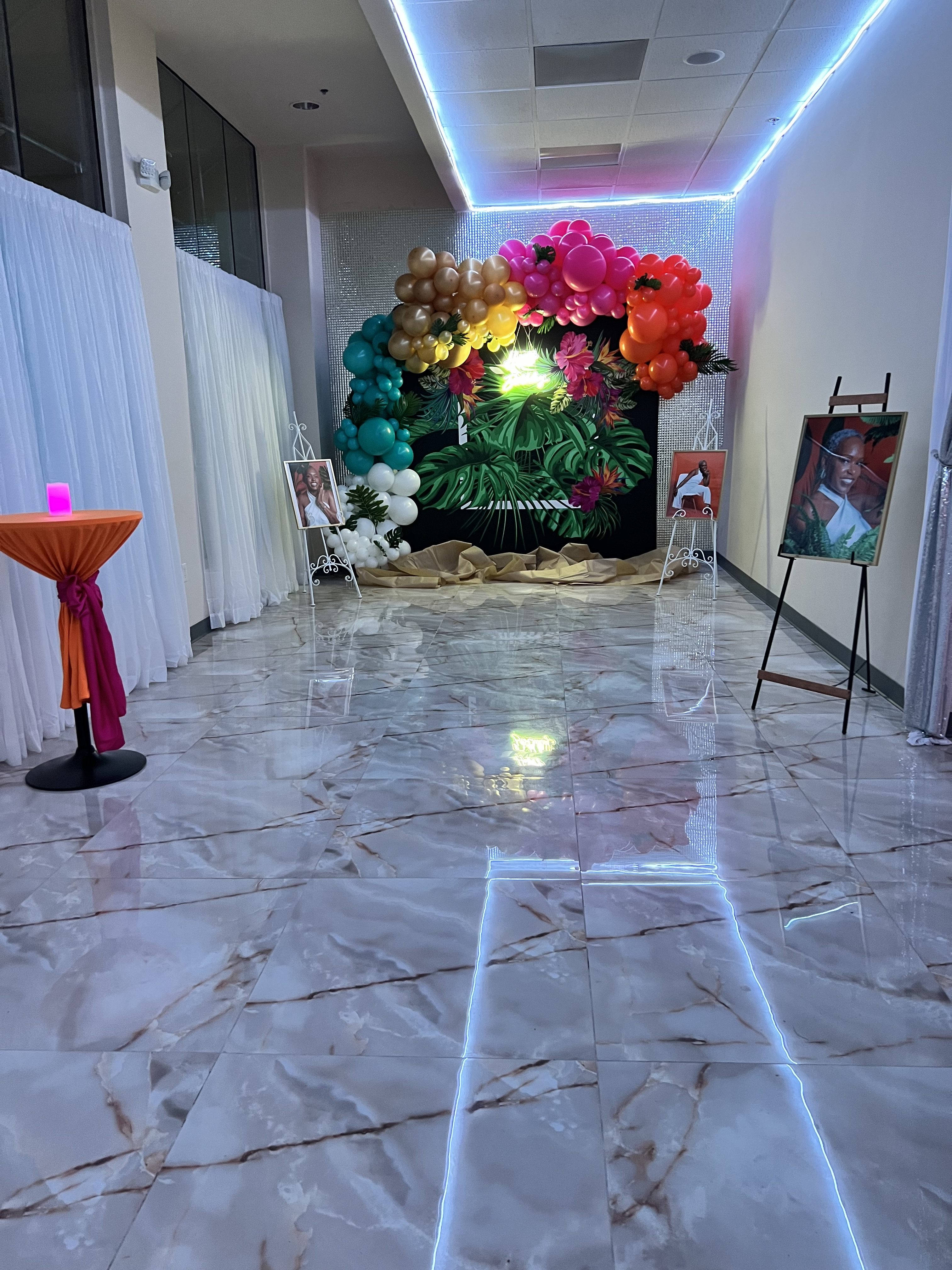 Decorative pathway at an event venue highlighted by led strips on the floor, leading to a vibrant tropical-themed balloon arch and photo displays on easels.