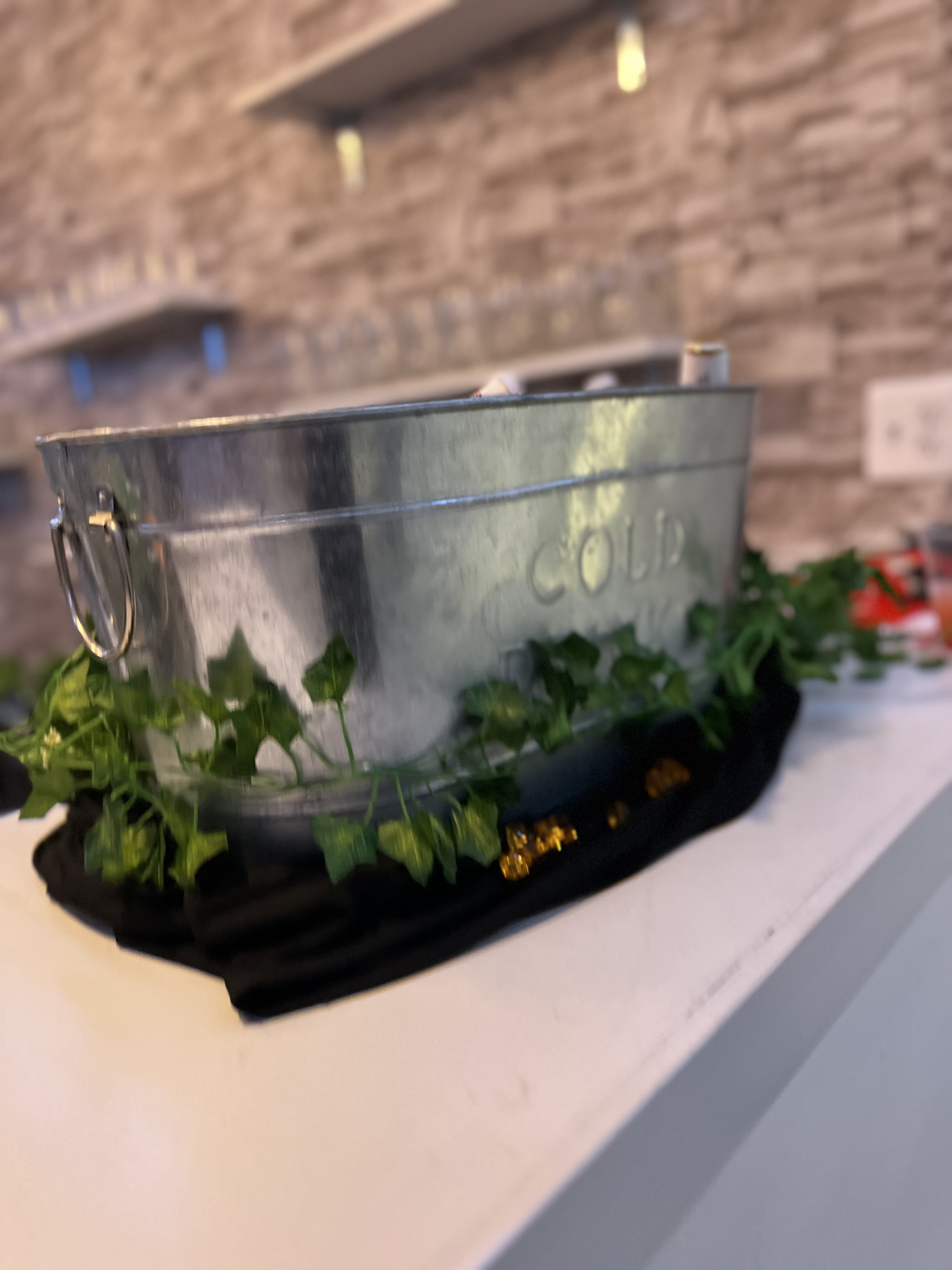 Metal container with the words "be cold" on a white mantel, adorned with green ivy and fairy lights, against a brick wall background.