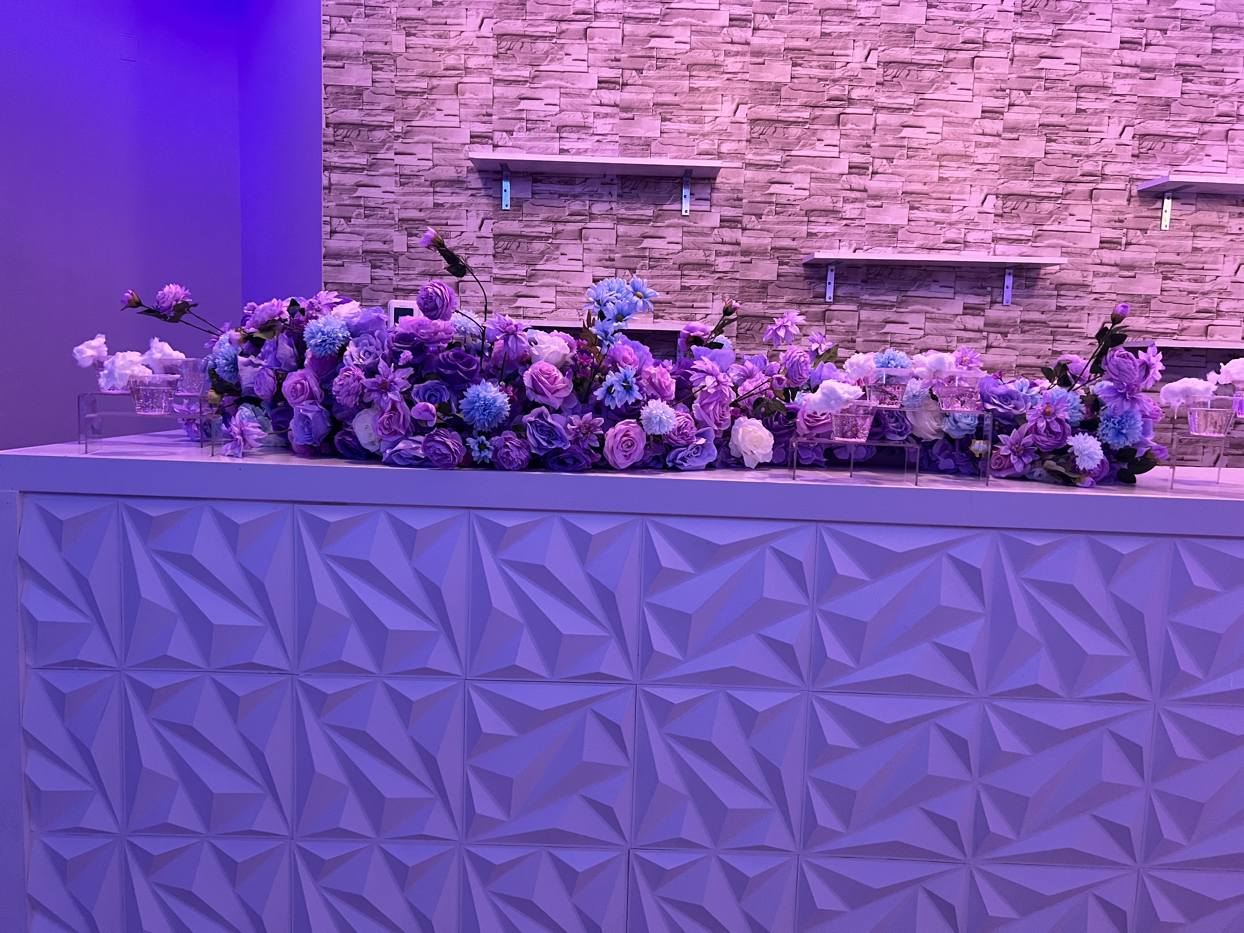 A beautifully arranged row of flowers in shades of blue, purple, and white on a white textured counter, against a stone wall lit by purple lighting.
