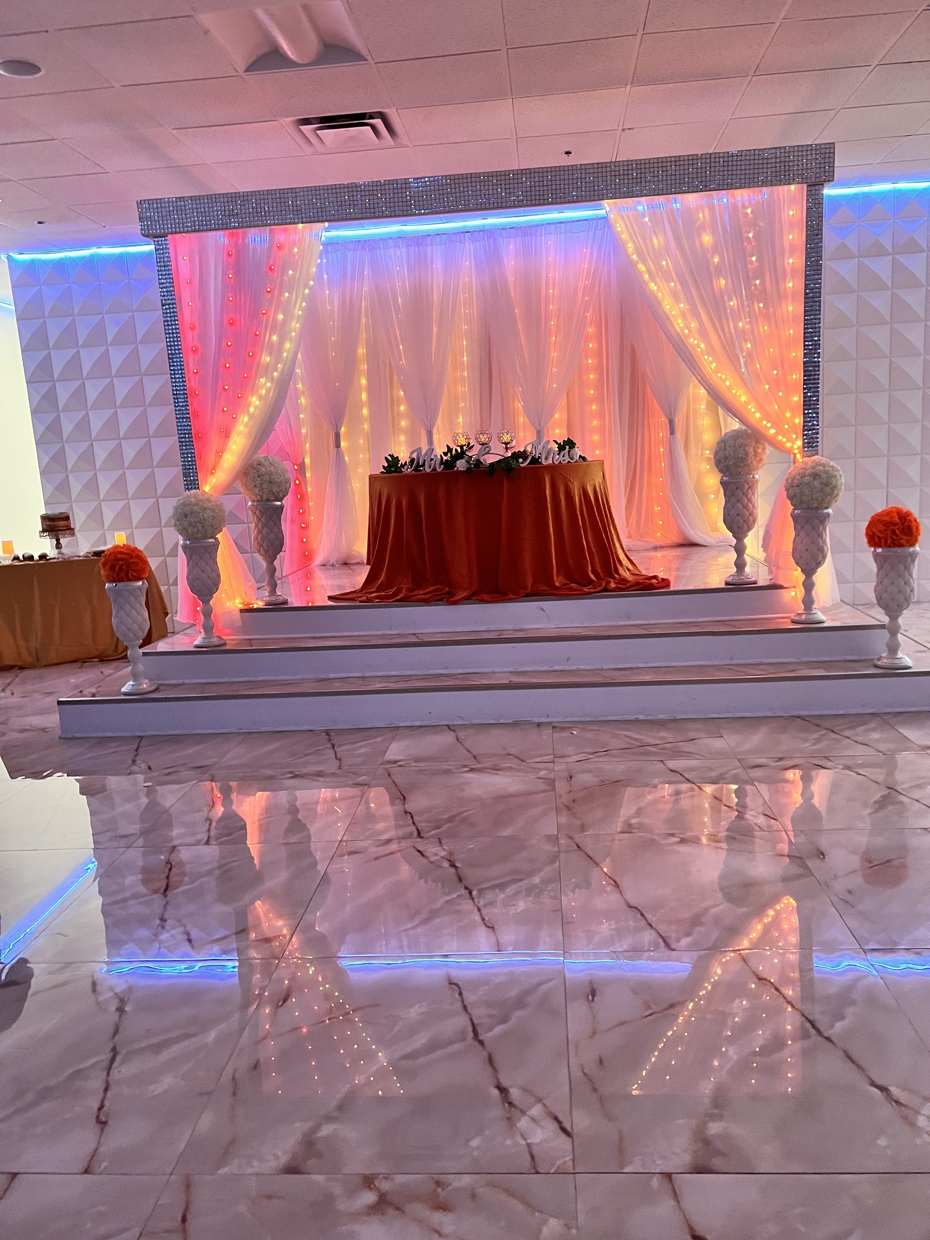 A wedding stage decorated with orange and white floral arrangements on pedestals, an elegant red tablecloth on the central table, and a backdrop of draped orange curtains with soft lighting.