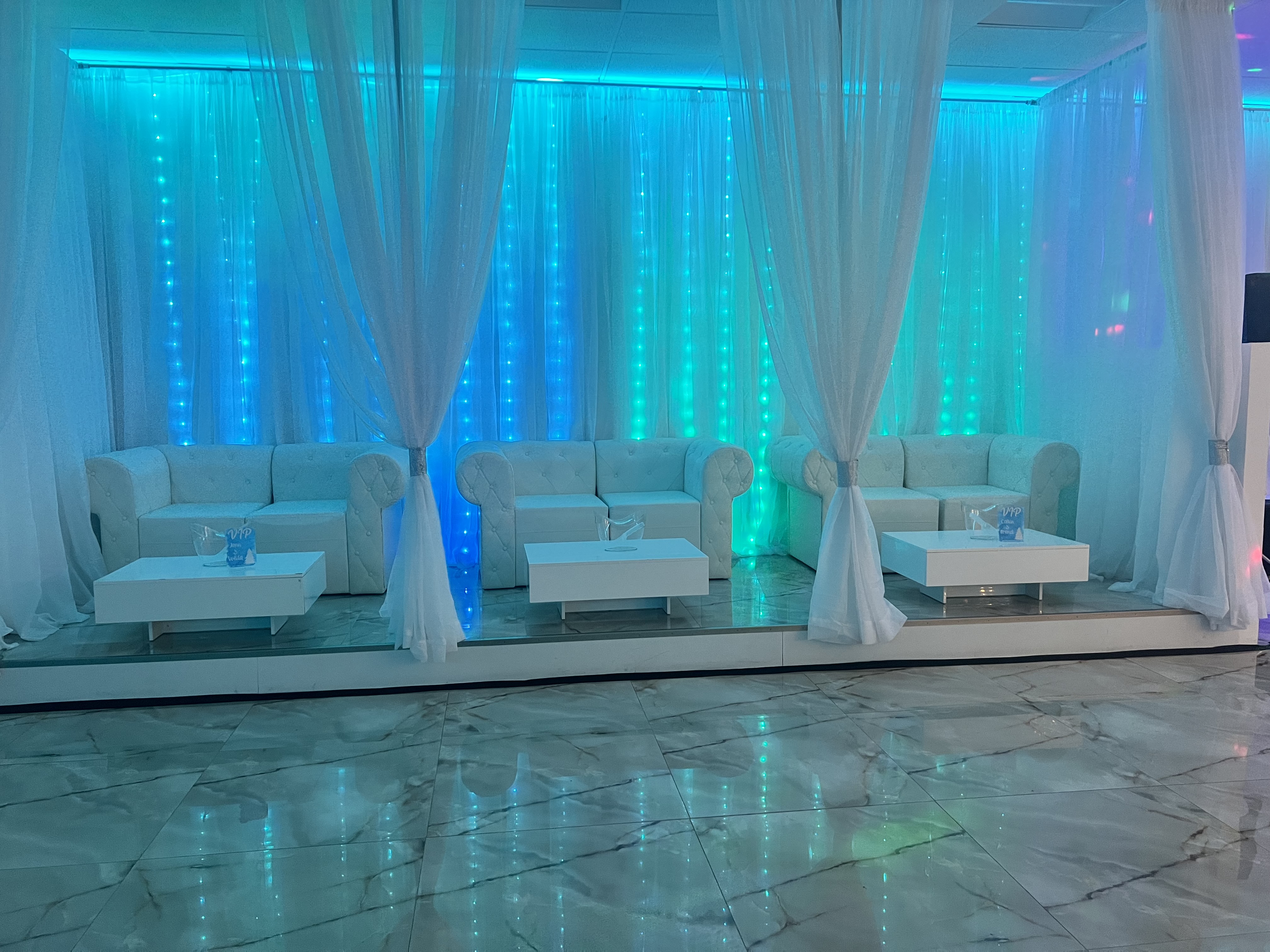 Elegant event space with a long white sofa and matching coffee tables on a marble floor, set against a backdrop of blue led-lighted curtains.
