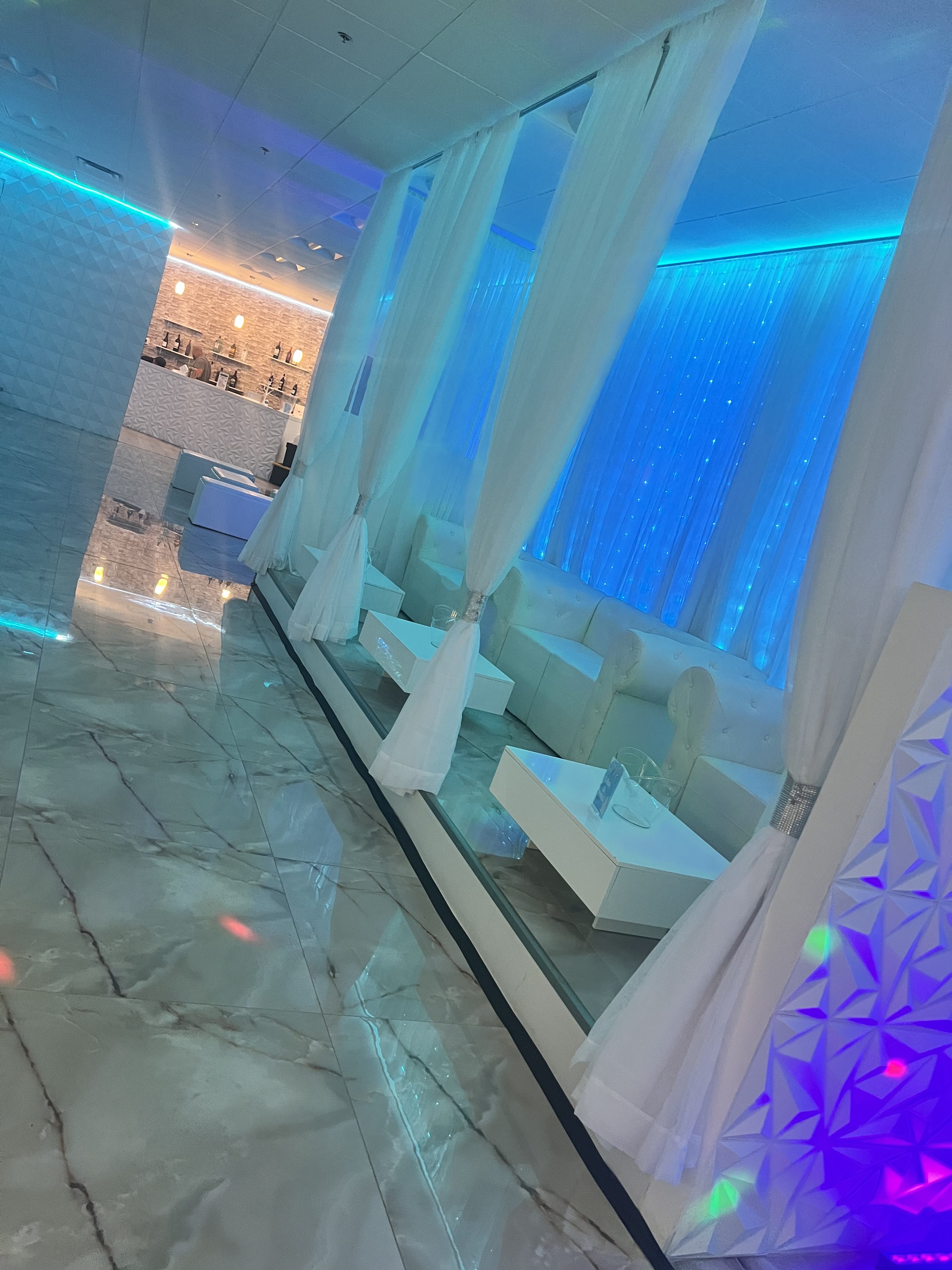 A modern lounge area with white furniture and curtains, illuminated by blue lights and a glowing bar in the background. the floor is glossy with reflective marble tiles.