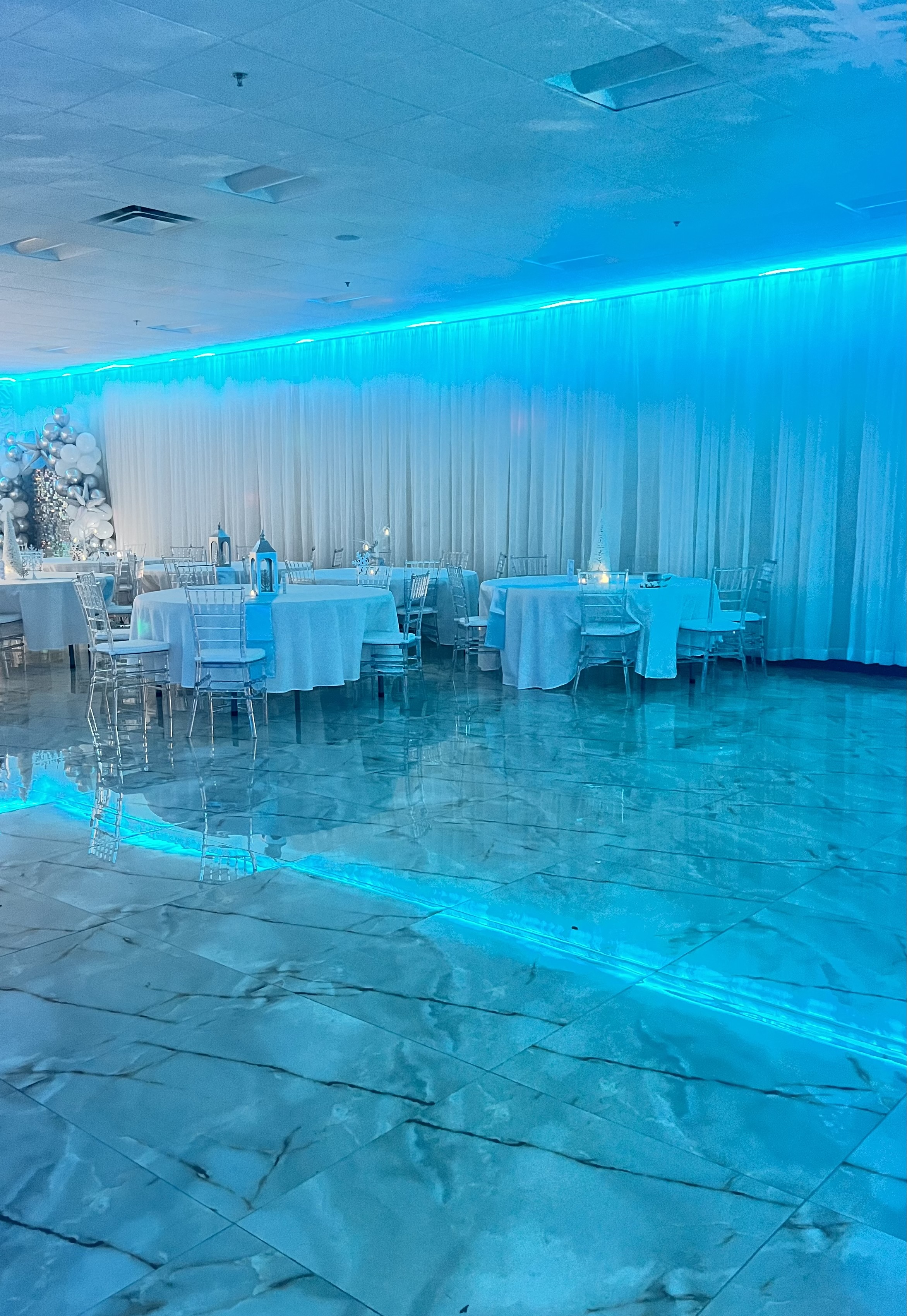 An elegant event hall with blue lighting, featuring tables draped with white linens, clear acrylic chairs, and a sleek, glossy floor.